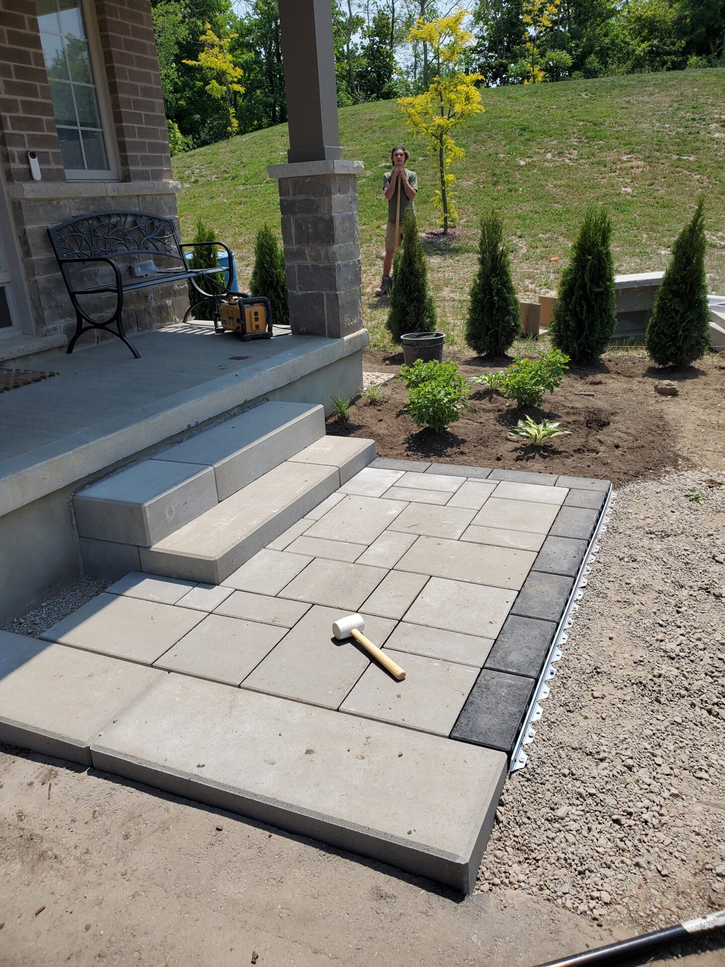 A newly constructed patio with a mallet, surrounded by shrubs. A person stands nearby, and a bench is on the porch.