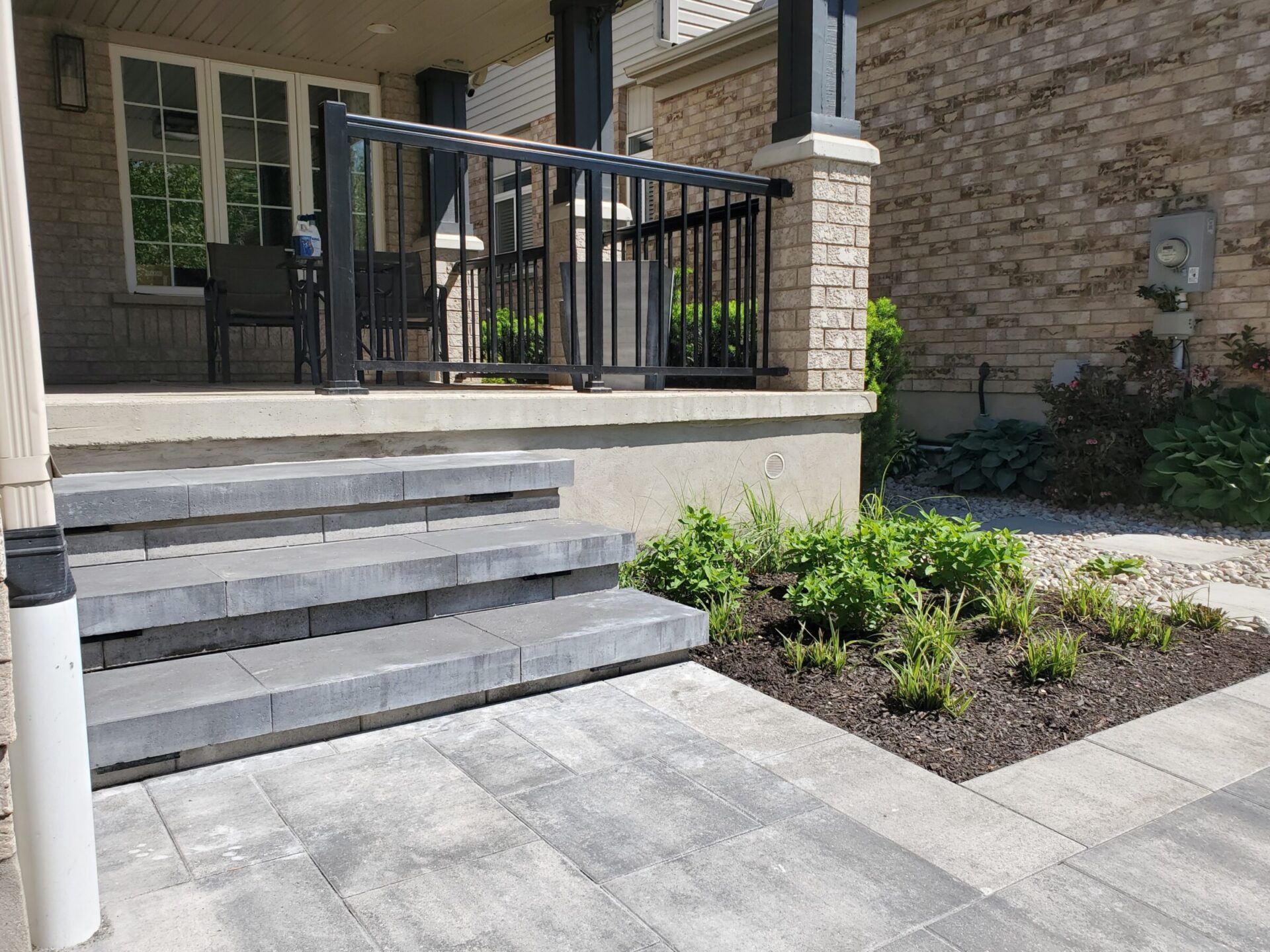 A brick house porch with steps, black railings, patio furniture, and a landscaped garden featuring mulch and various plants.