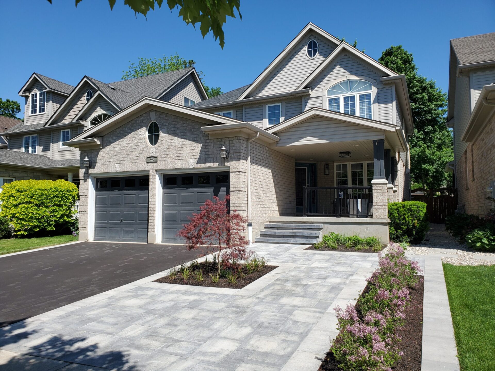 Two-story suburban house with gray siding, two-car garage, and landscaped front yard. Clear blue sky and lush greenery surround the home.