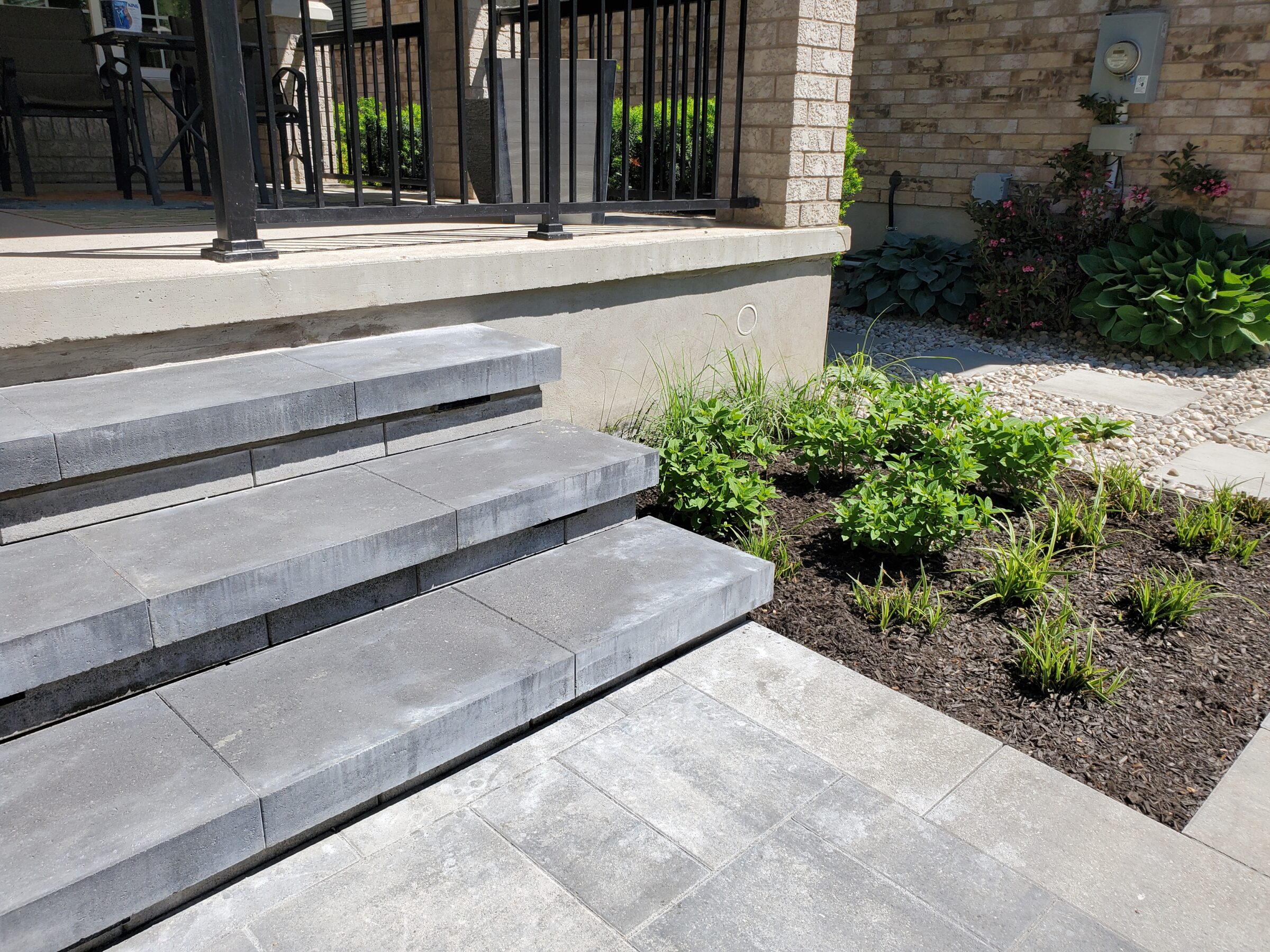 Stone steps lead to a porch with black railing and a garden featuring green plants and mulch, next to a brick wall.