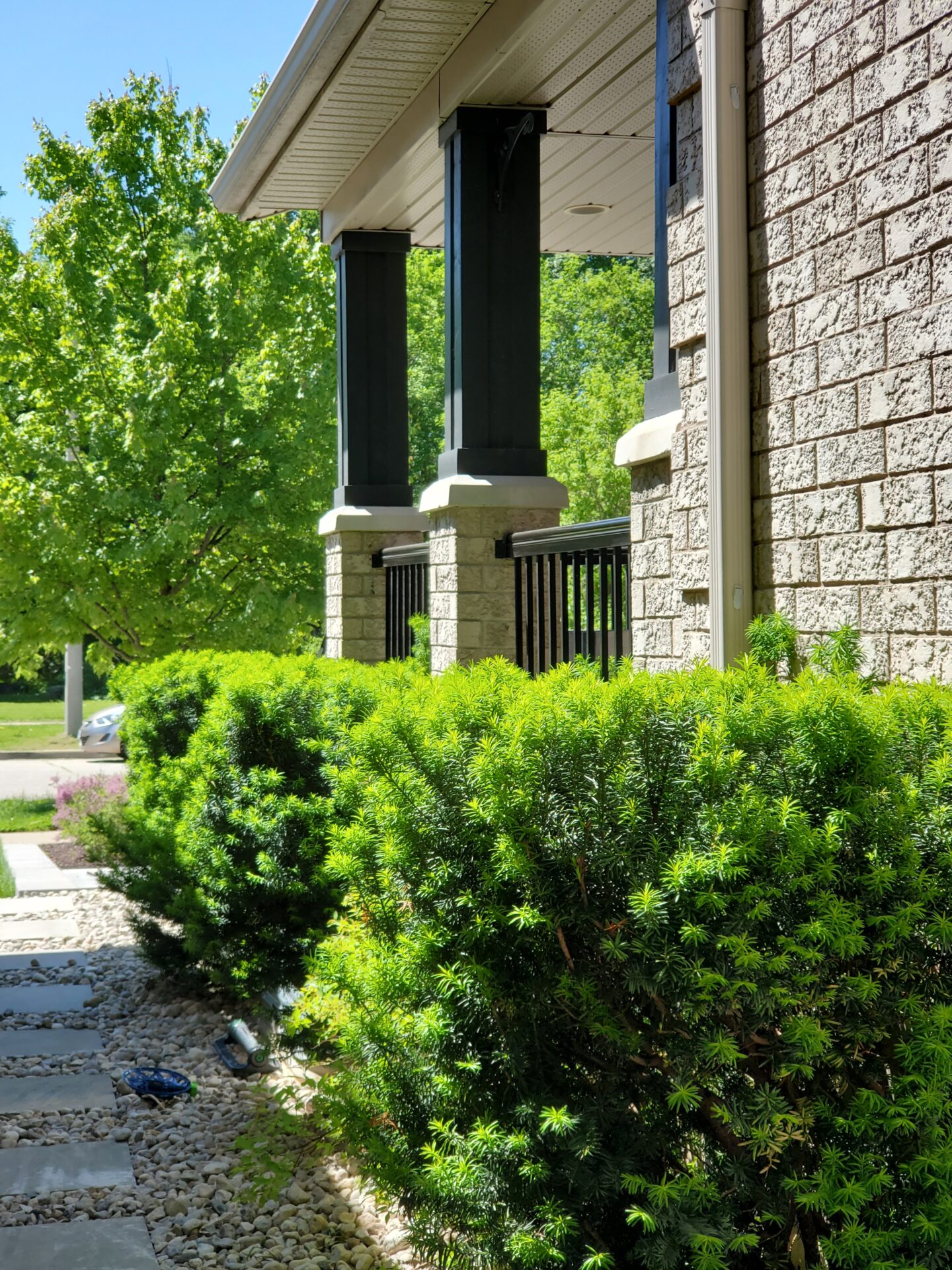 A modern porch with black pillars and brickwork, lush green shrubs, and a stone path in a residential neighborhood setting.