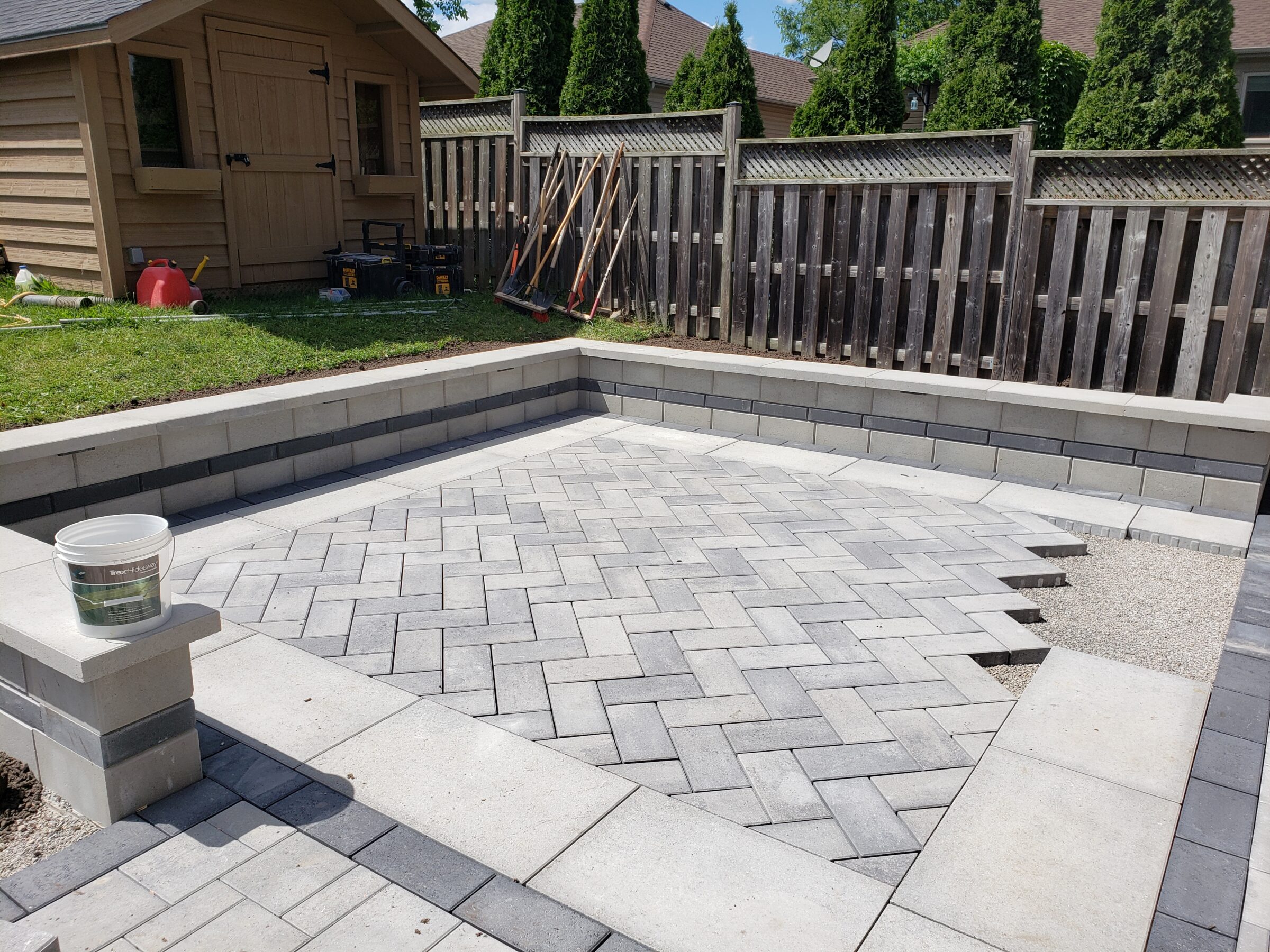 A backyard with partially completed stone patio, surrounded by a wooden fence. A shed and garden tools are visible in the background.