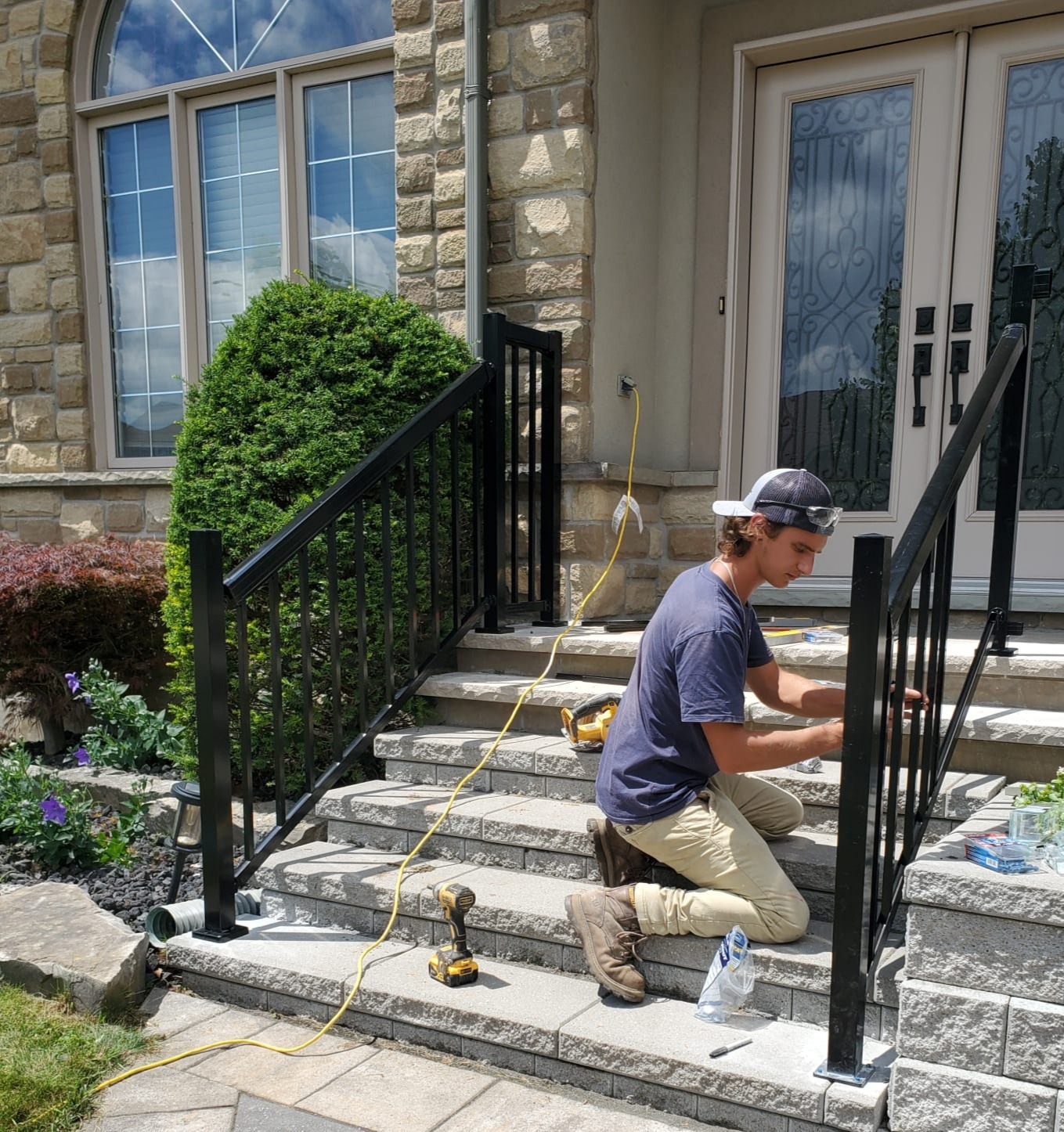 A person is installing a black metal railing on stone steps. Tools, greenery, and a brick house facade are visible.