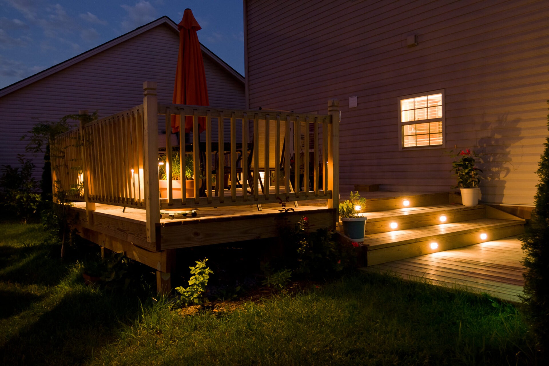 A cozy, illuminated backyard deck with plants, a table, chairs, and an umbrella against a night sky. Warm, inviting atmosphere enhanced by step lights.