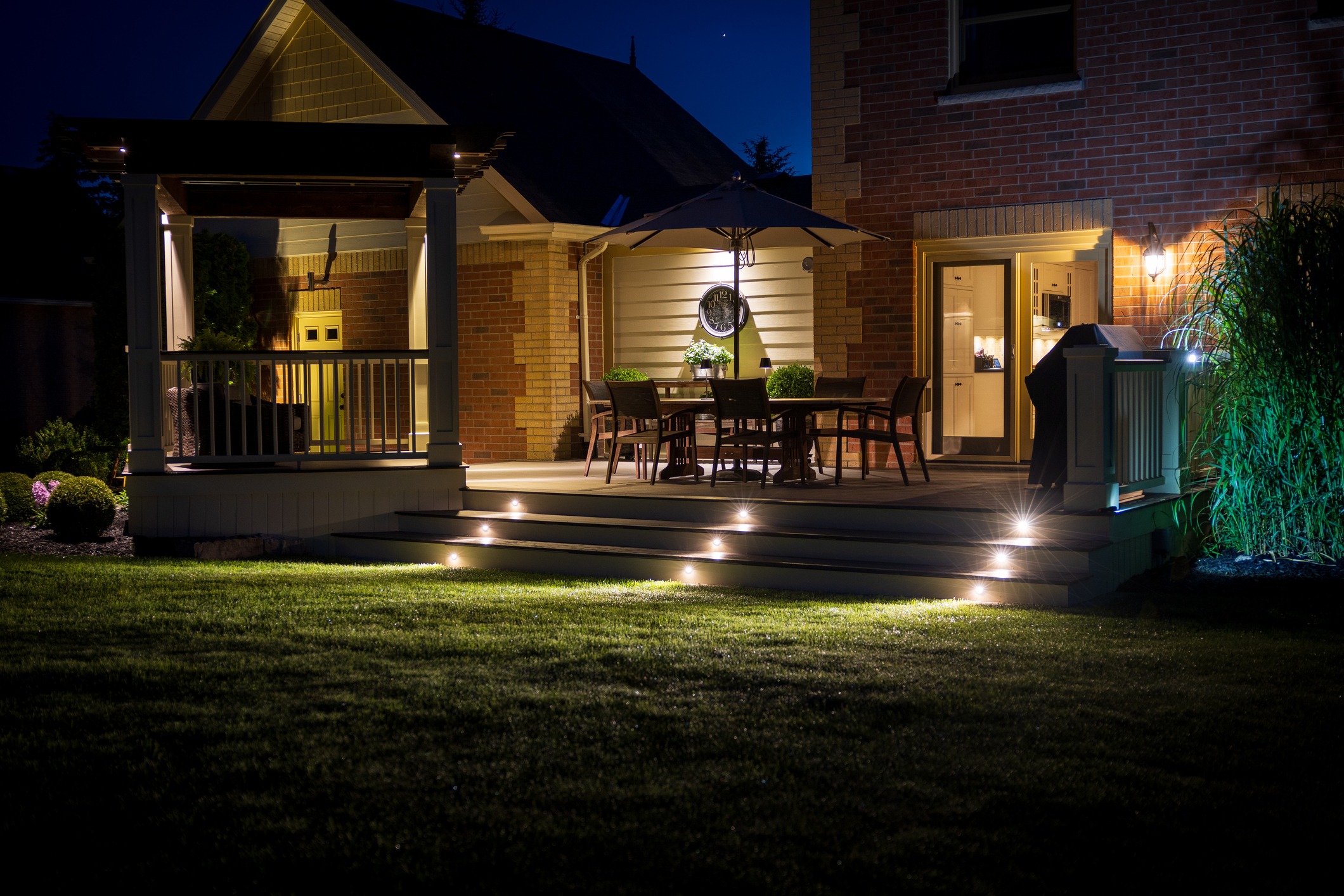 A cozy, illuminated patio at night, featuring outdoor seating and a pergola, attached to a brick house with a well-maintained lawn.