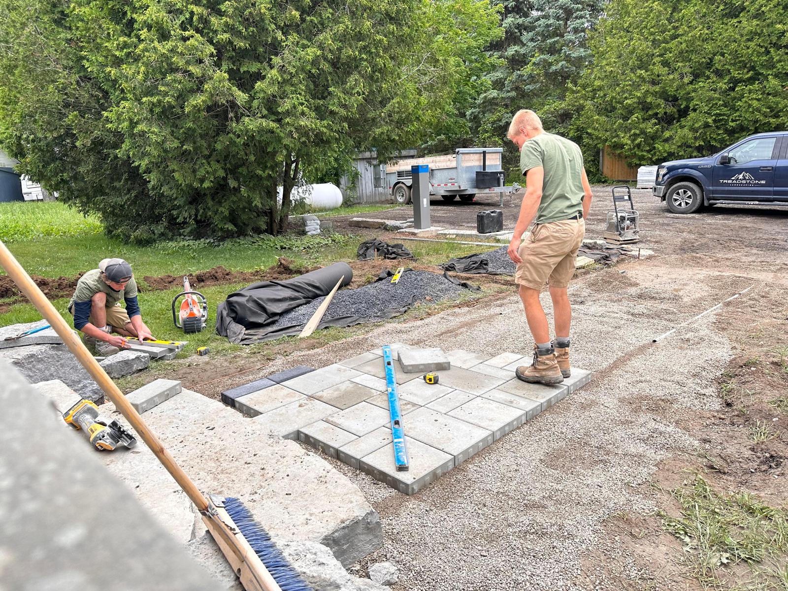 Two people working on a stone pathway in a garden, surrounded by tools and equipment, with nearby trees and parked vehicles in the background.