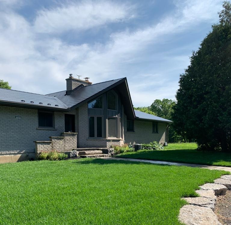 A modern brick house with large windows is surrounded by green grass and trees under a partly cloudy sky.