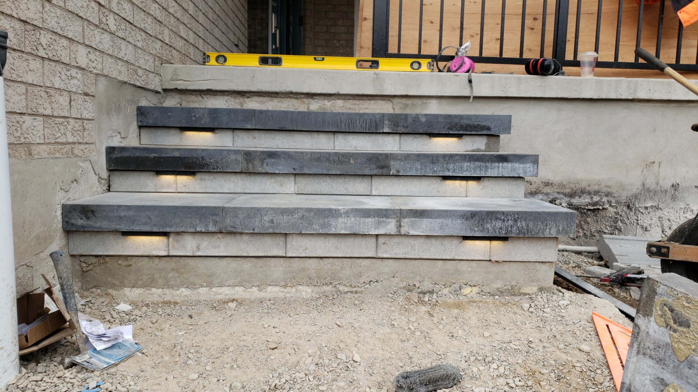 Stone steps under construction with embedded lights. Tools like a level and shovel are scattered around; a railing is partially visible.