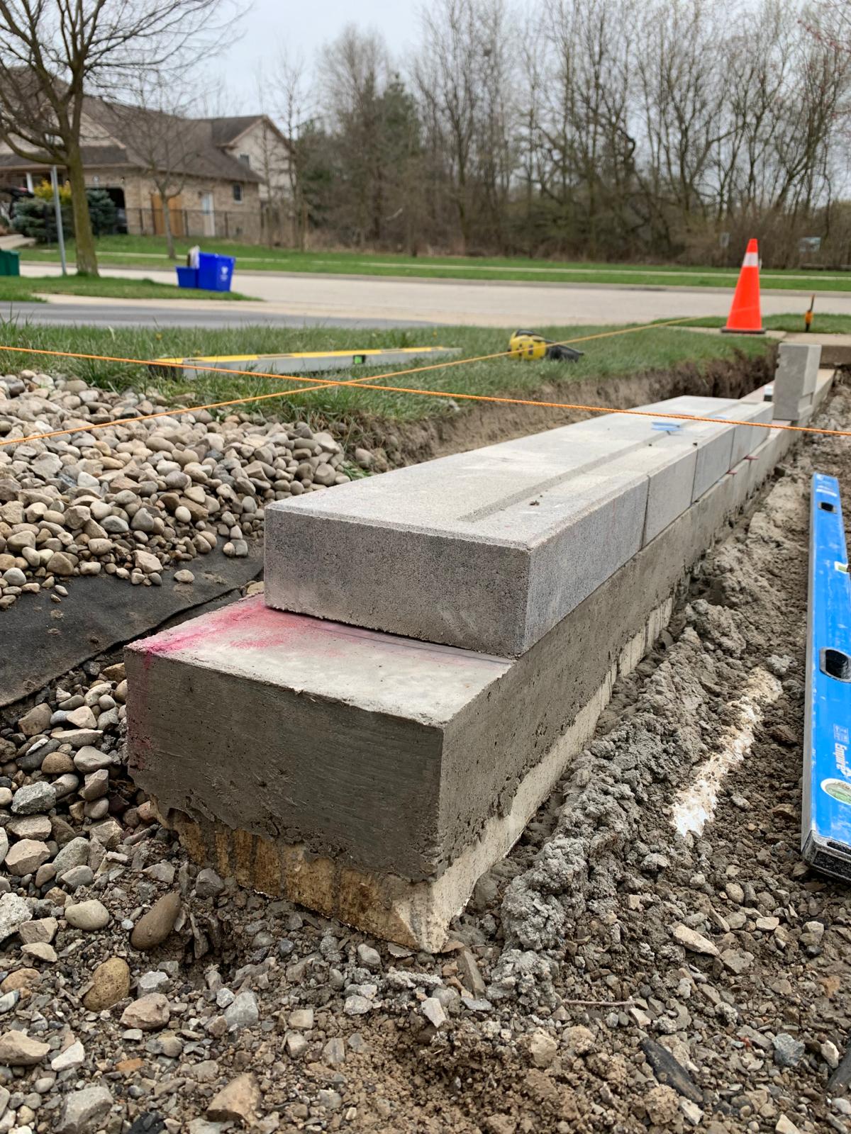 Concrete blocks and bricks under construction near a street with trees and a house in the background. Nearby orange cone and tools visible.