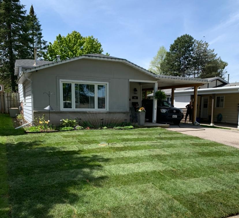 A single-story house with a fresh lawn, carport, and car. A person stands near the car beneath a clear blue sky.