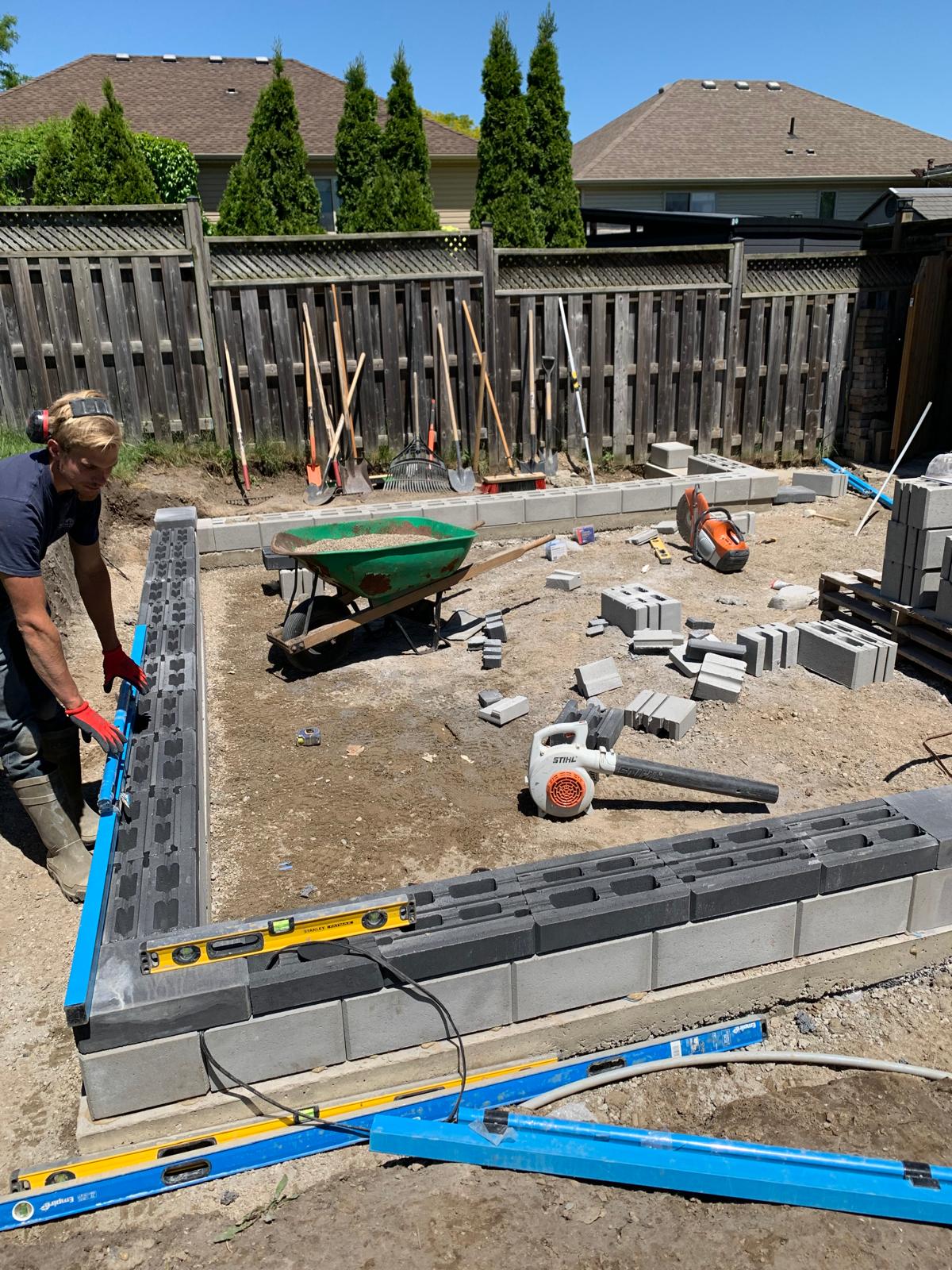 A person is building a low cinder block wall in a yard with tools, a wheelbarrow, and a wooden fence.
