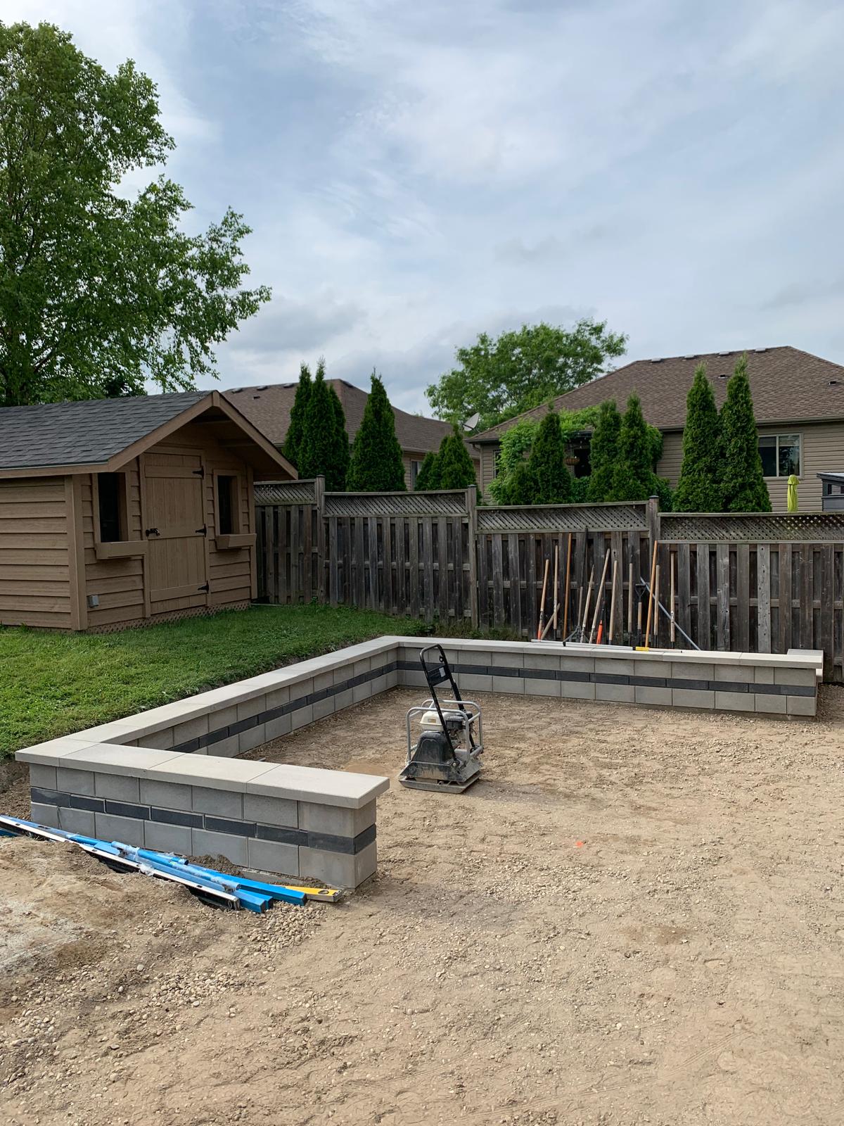 A backyard construction site features a low brick wall, small wooden shed, and a fenced area with tools and landscaping materials visible.