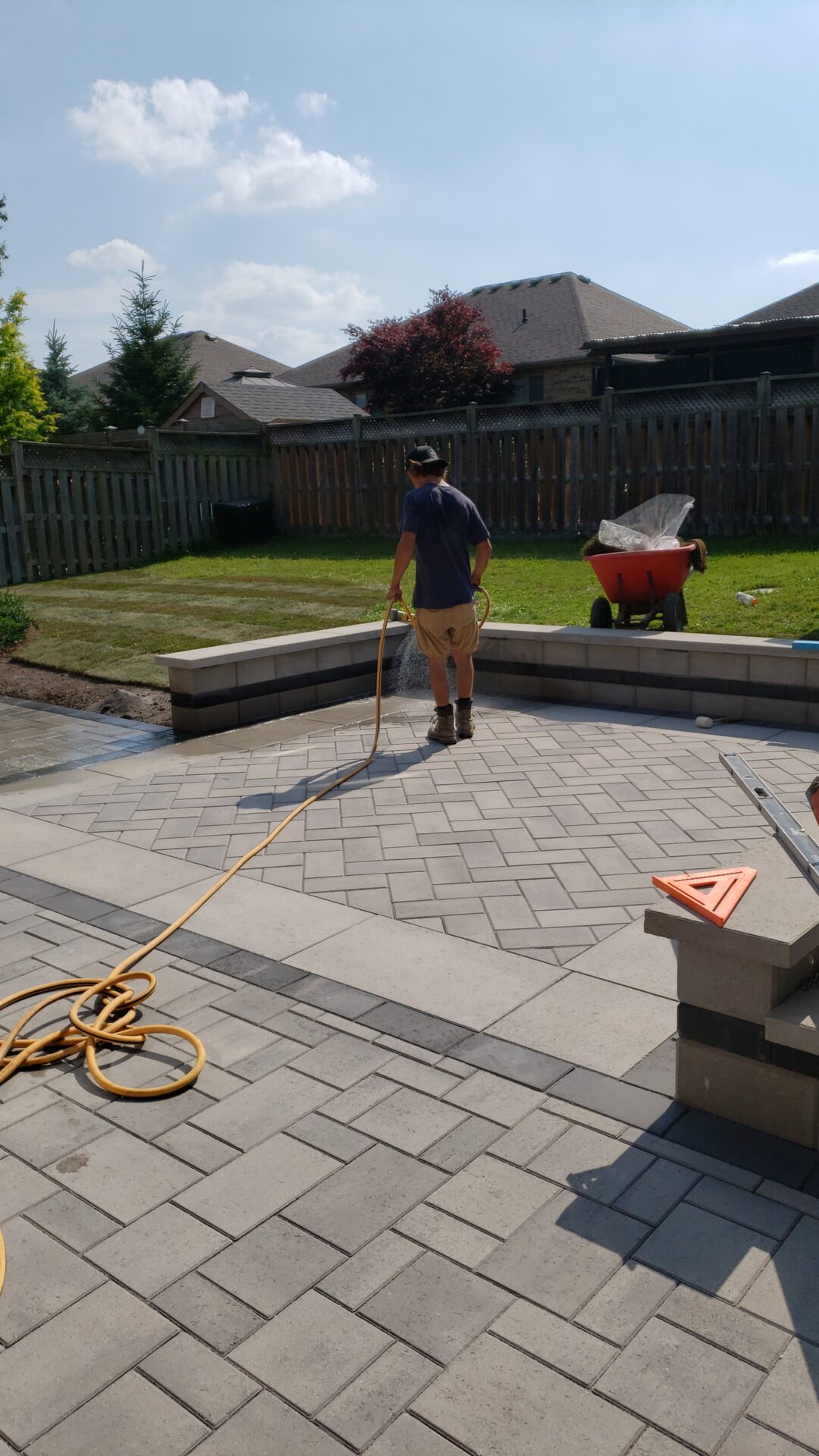 A person is watering newly installed patio stones in a backyard under a sunny sky, with a wheelbarrow and fence in the background.