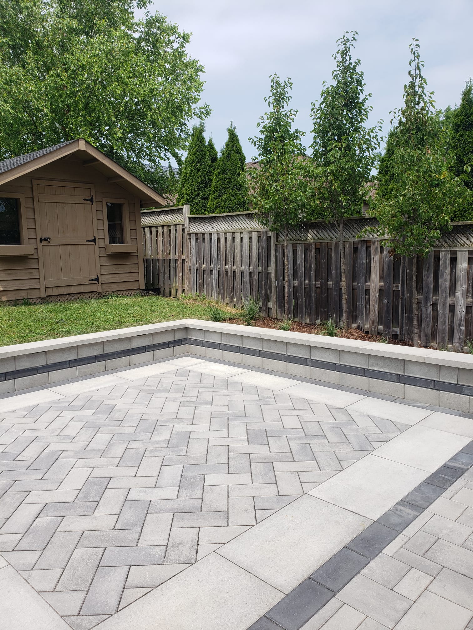 A garden scene with a wooden shed, fenced border, new paved patio, and green trees under a clear sky. No people are visible.