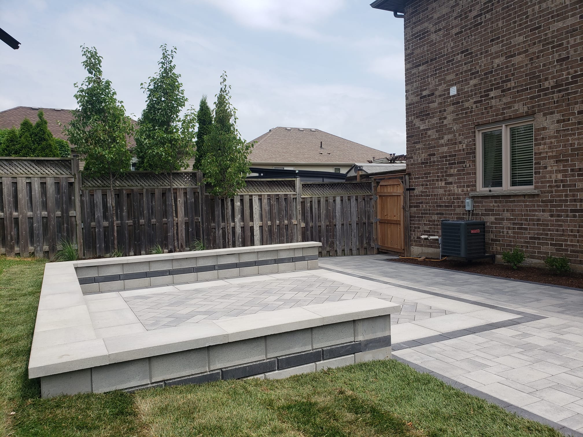 A modern backyard features a sleek stone patio and wall, bordered by wooden fencing and young trees. Brick house visible on the right.