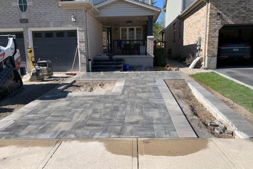 A house with a partially completed driveway is visible. Construction equipment and vehicles are nearby, with brickwork and landscaping in progress.