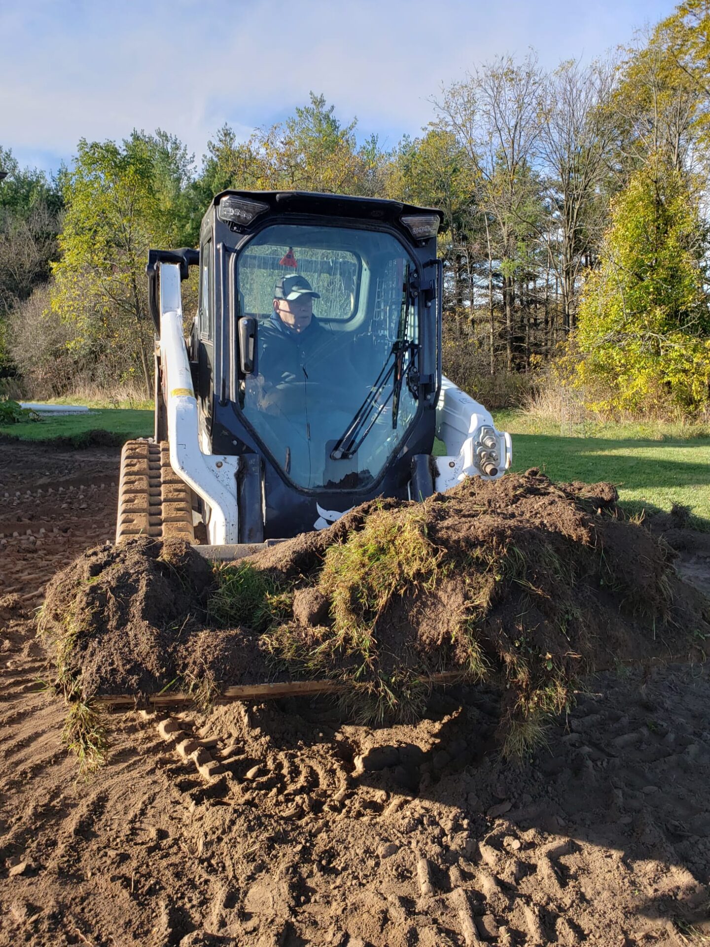 A person operates a skid steer loader filled with soil and grass, set against a backdrop of trees under a clear sky.