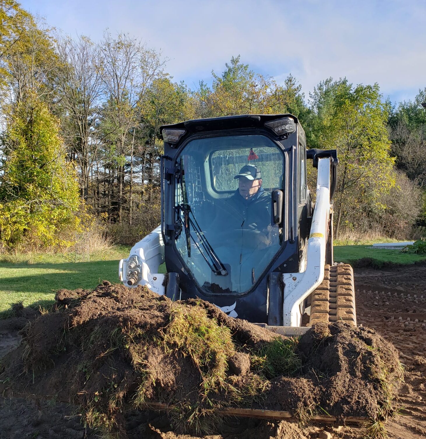 A person operates a skid steer loader filled with soil and grass, set against a backdrop of trees under a clear sky.