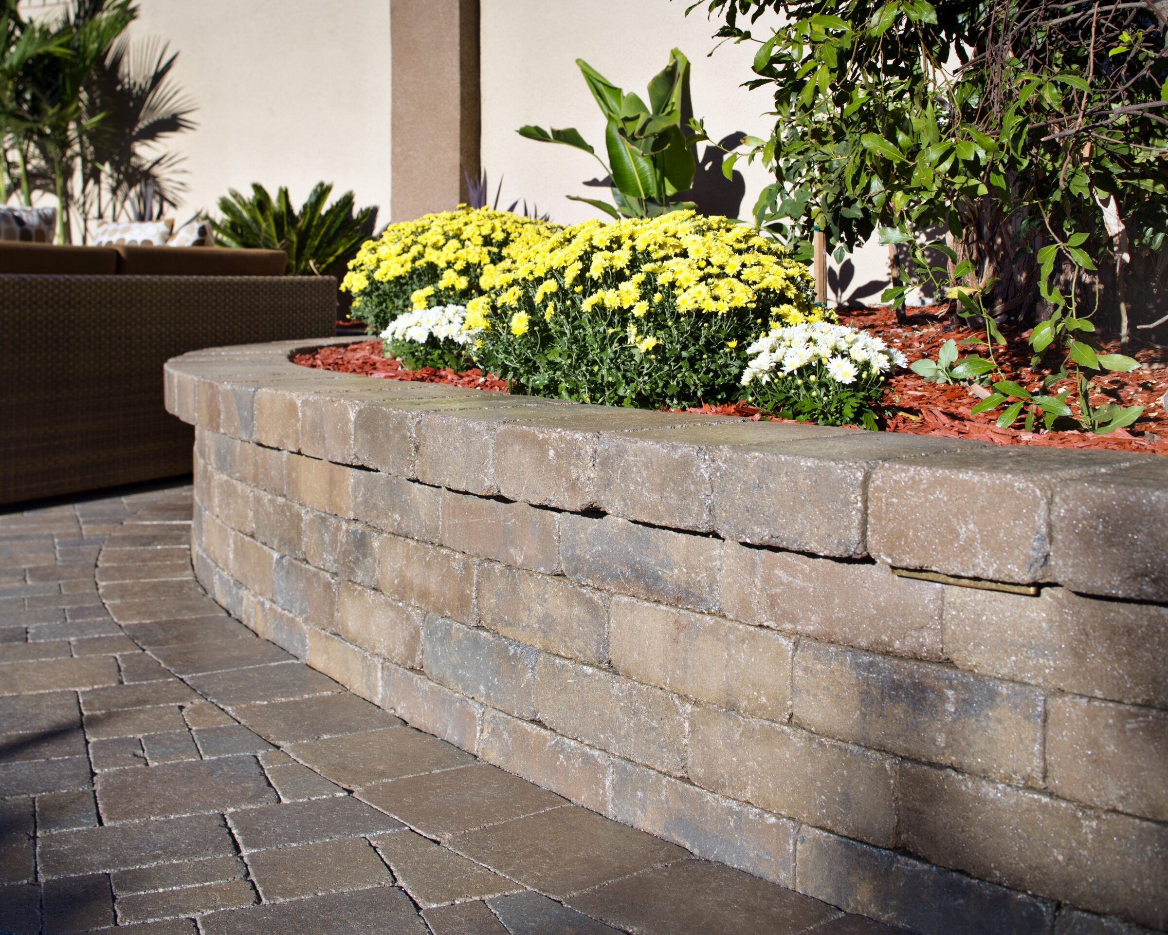 Decorative stone wall with vibrant yellow and white flowers, surrounded by lush greenery, accentuating a stylish outdoor patio with brick flooring.