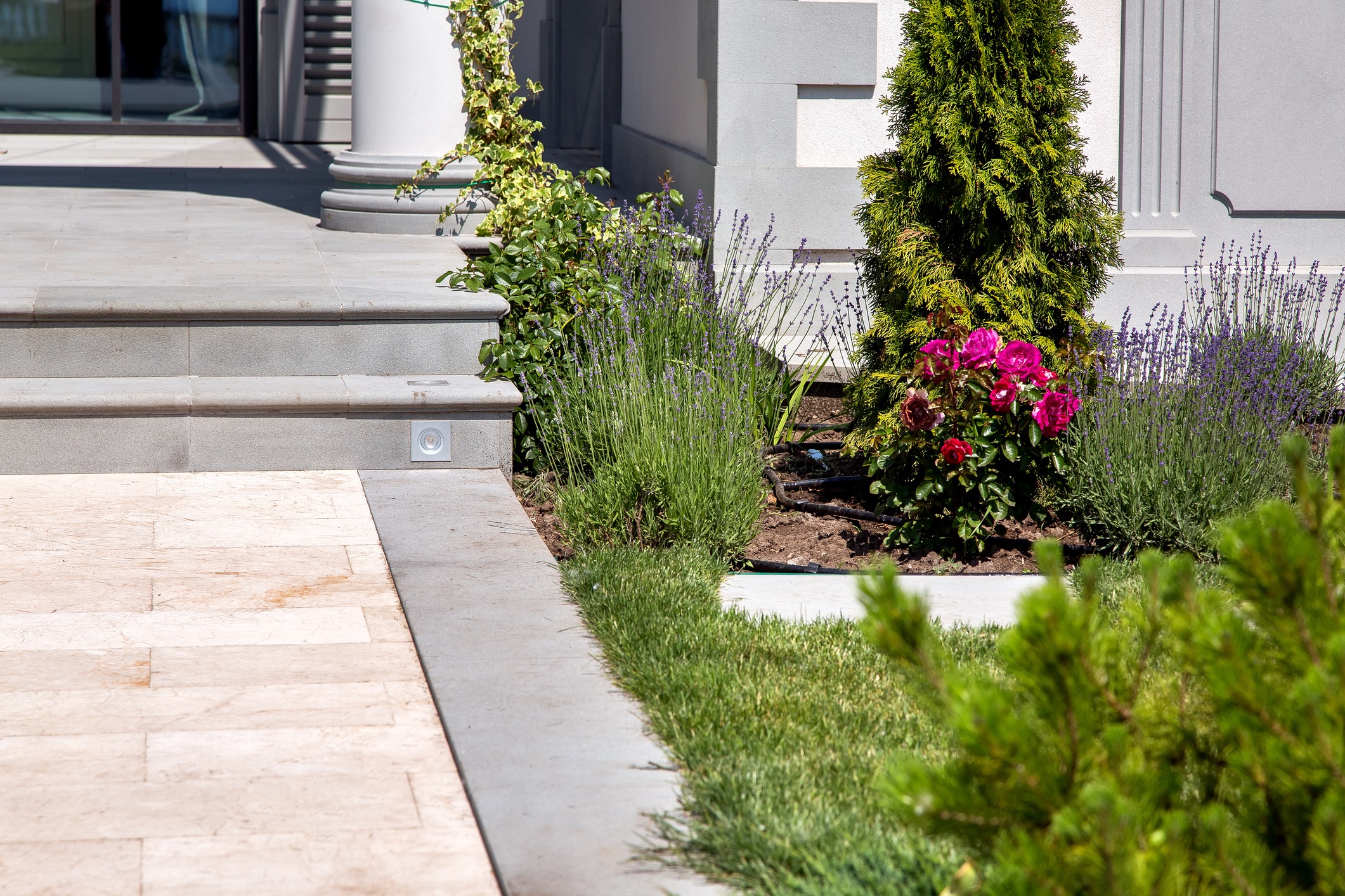 Ornate garden with lush greenery and blooming flowers surrounds elegant stone steps leading to a building entrance, accentuated by a tall column.