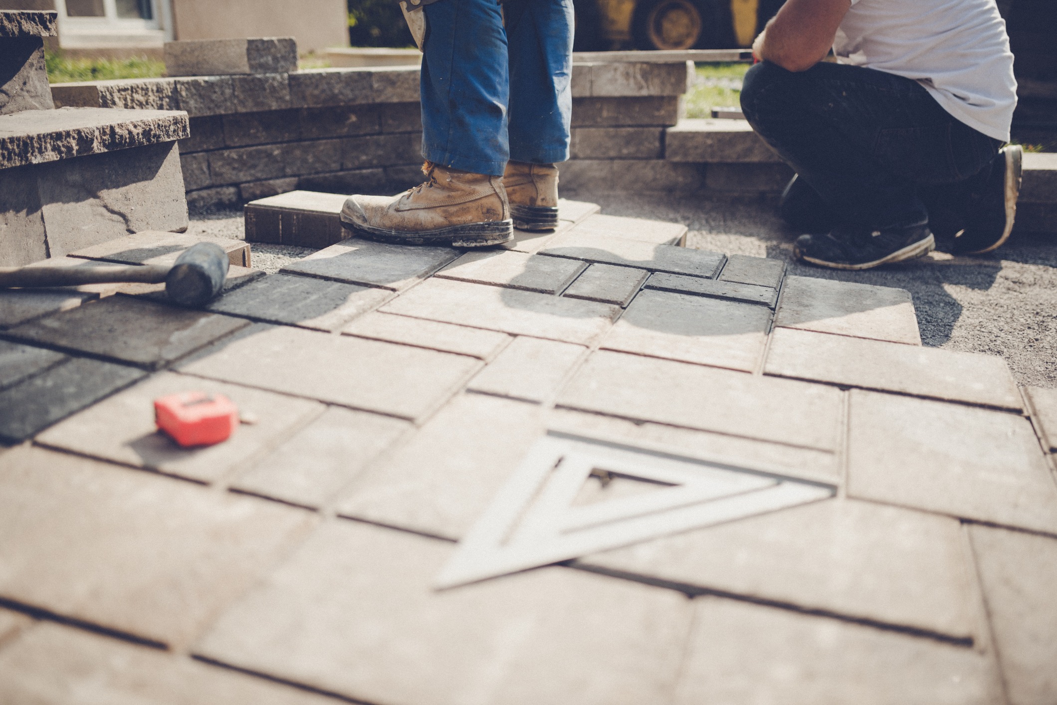 Two people working on a patio, surrounded by paving stones and tools, including a hammer and measuring tape. Construction in progress.