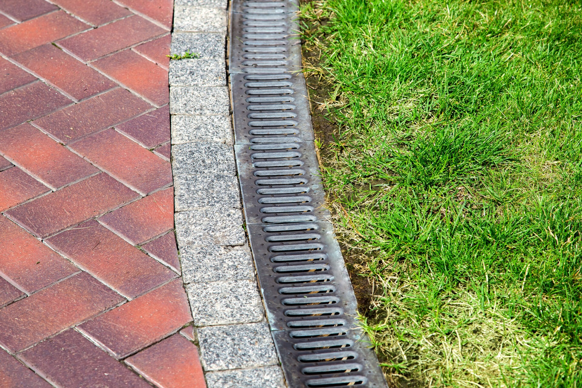 Red brick path with stone border alongside a metal drainage grate and lush green grass, capturing urban landscaping details.