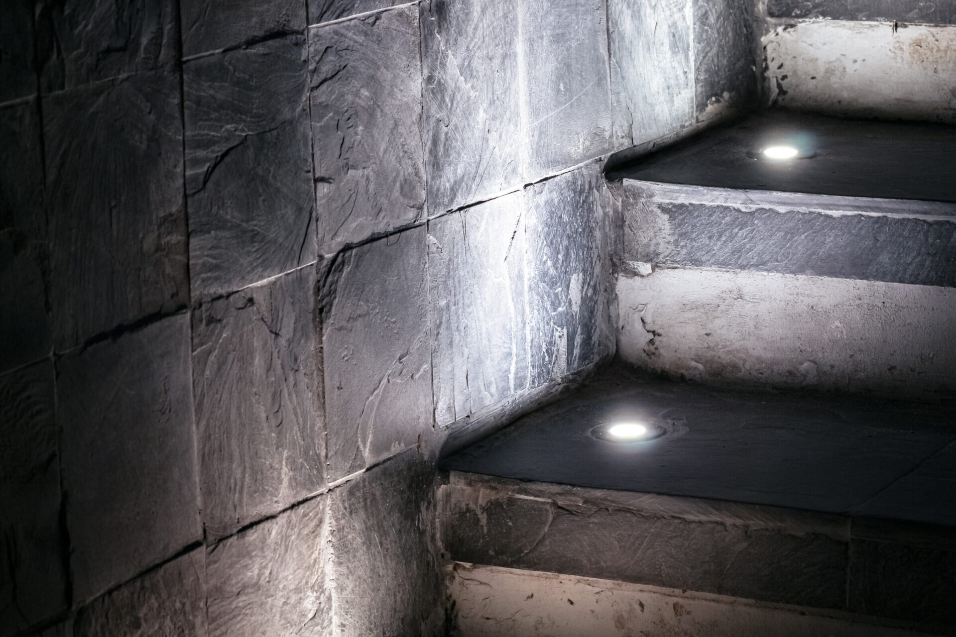 Dimly lit stone staircase with embedded floor lights, showcasing textured walls in a modern architectural setting, emphasizing a serene and minimalistic design.