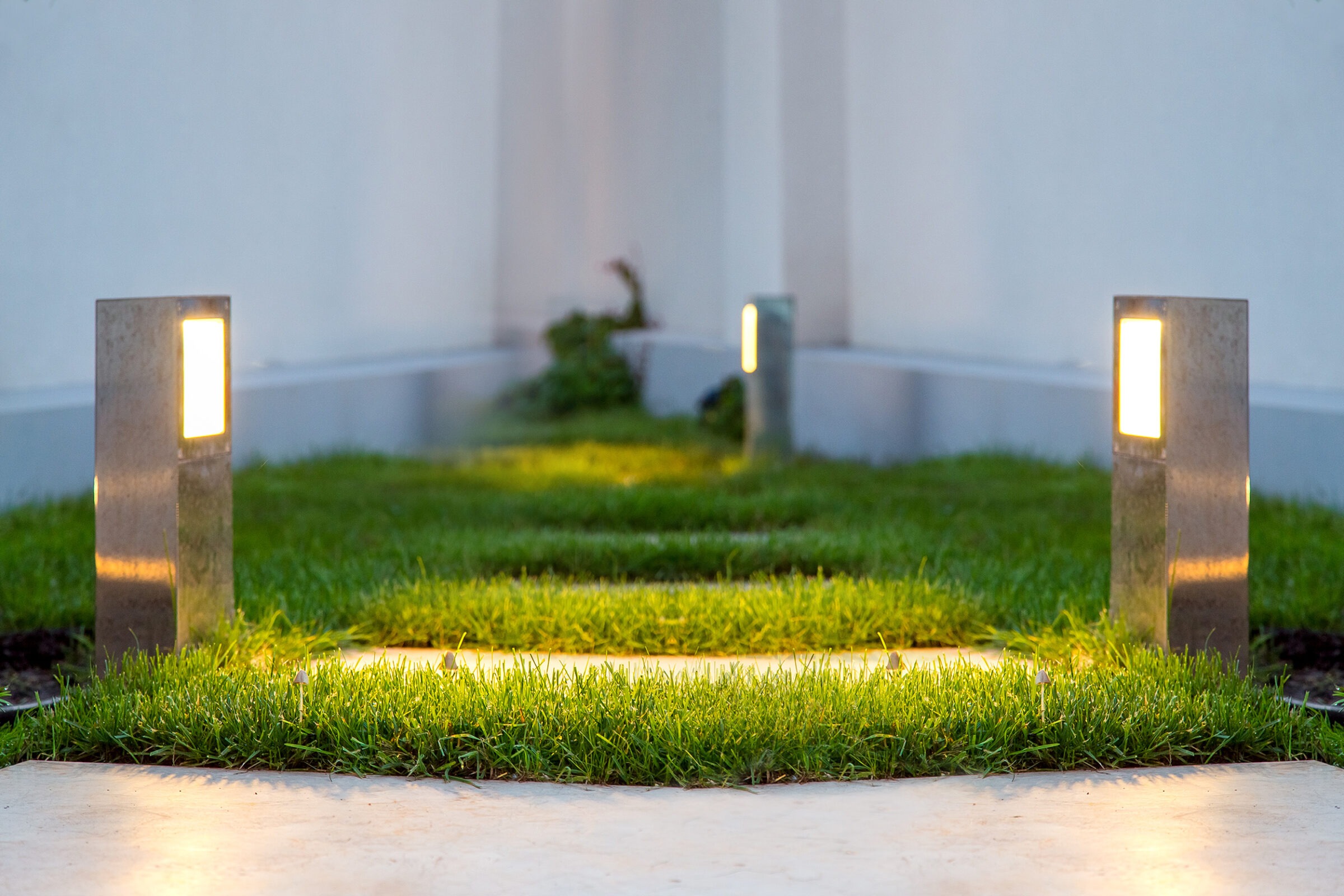 A minimalist garden path with modern lighting fixtures, lush green grass, and concrete surfaces, framed by a clean, white wall backdrop.
