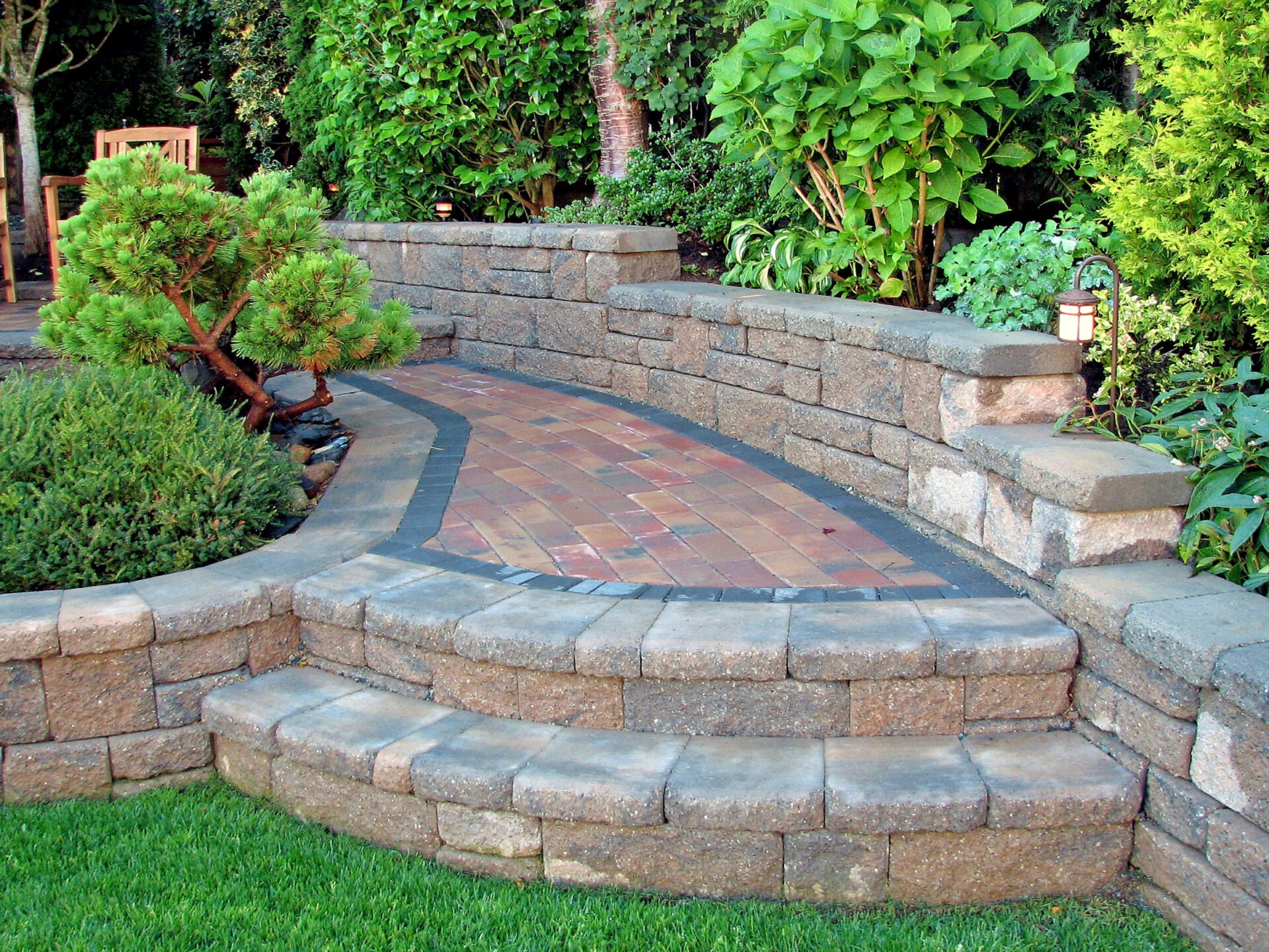 A serene garden features a stone pathway with steps, bordered by lush green plants and trees in a peaceful outdoor setting.