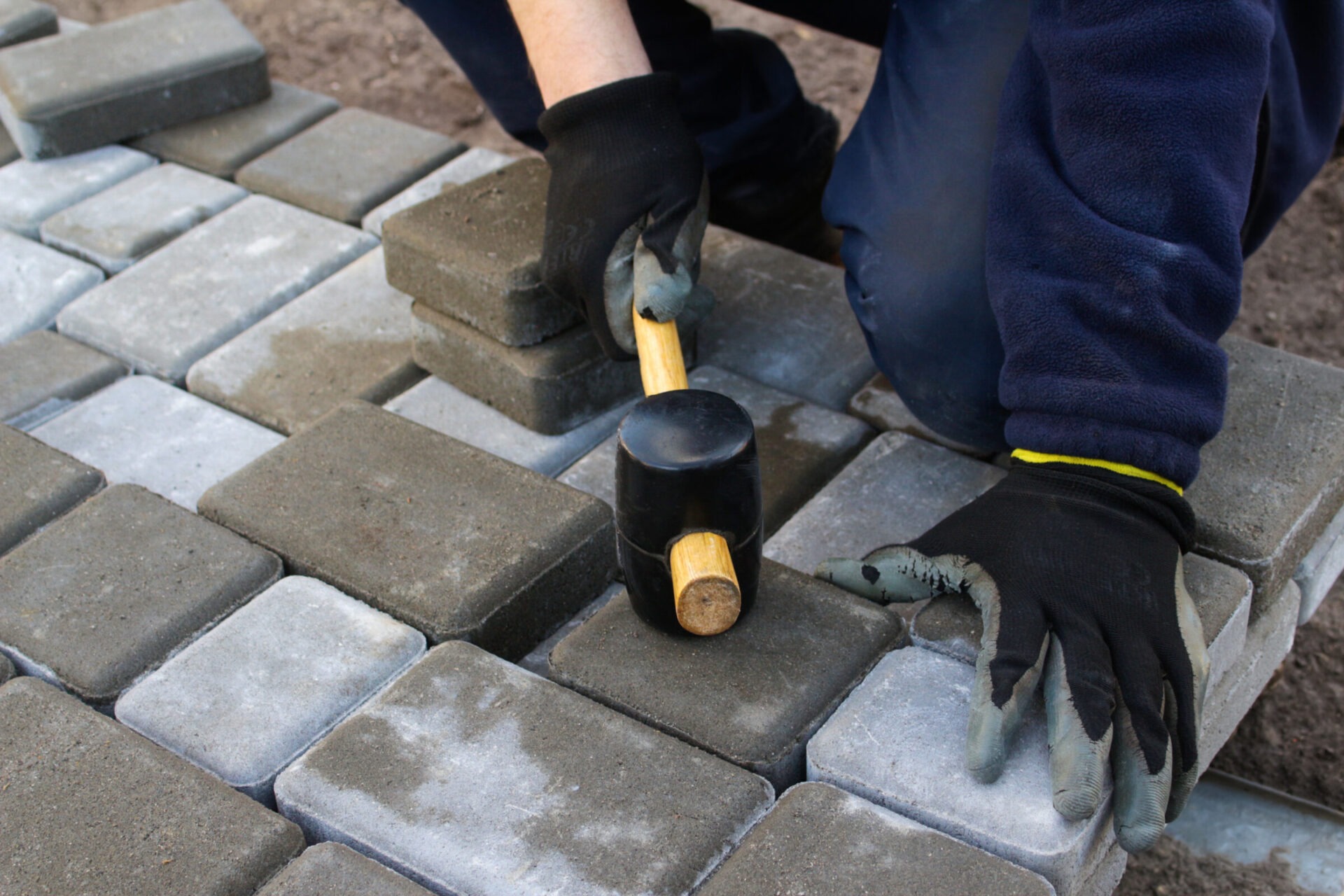 A person wearing gloves is using a mallet to lay gray and beige paving stones on the ground, focusing on alignment and placement.