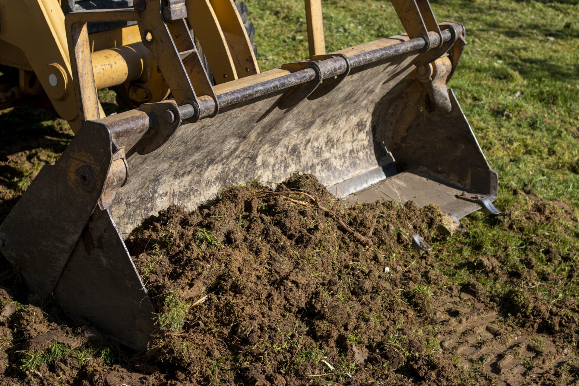 A bulldozer blade pushes a pile of dirt and grass on a sunny day at a construction site with green grass.