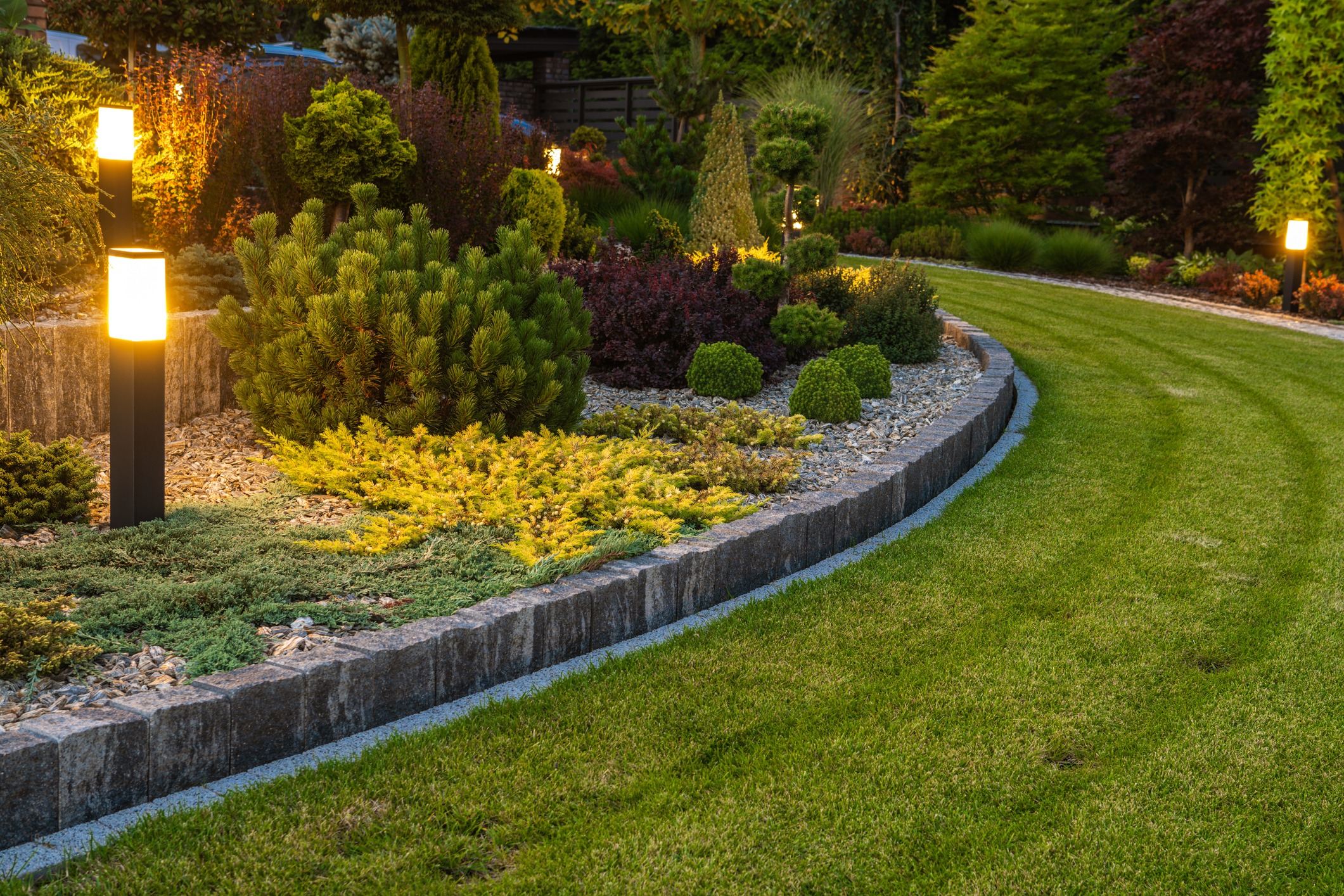 A beautifully landscaped garden with neatly trimmed grass, vibrant bushes, curved stone border, and glowing pathway lights at dusk.