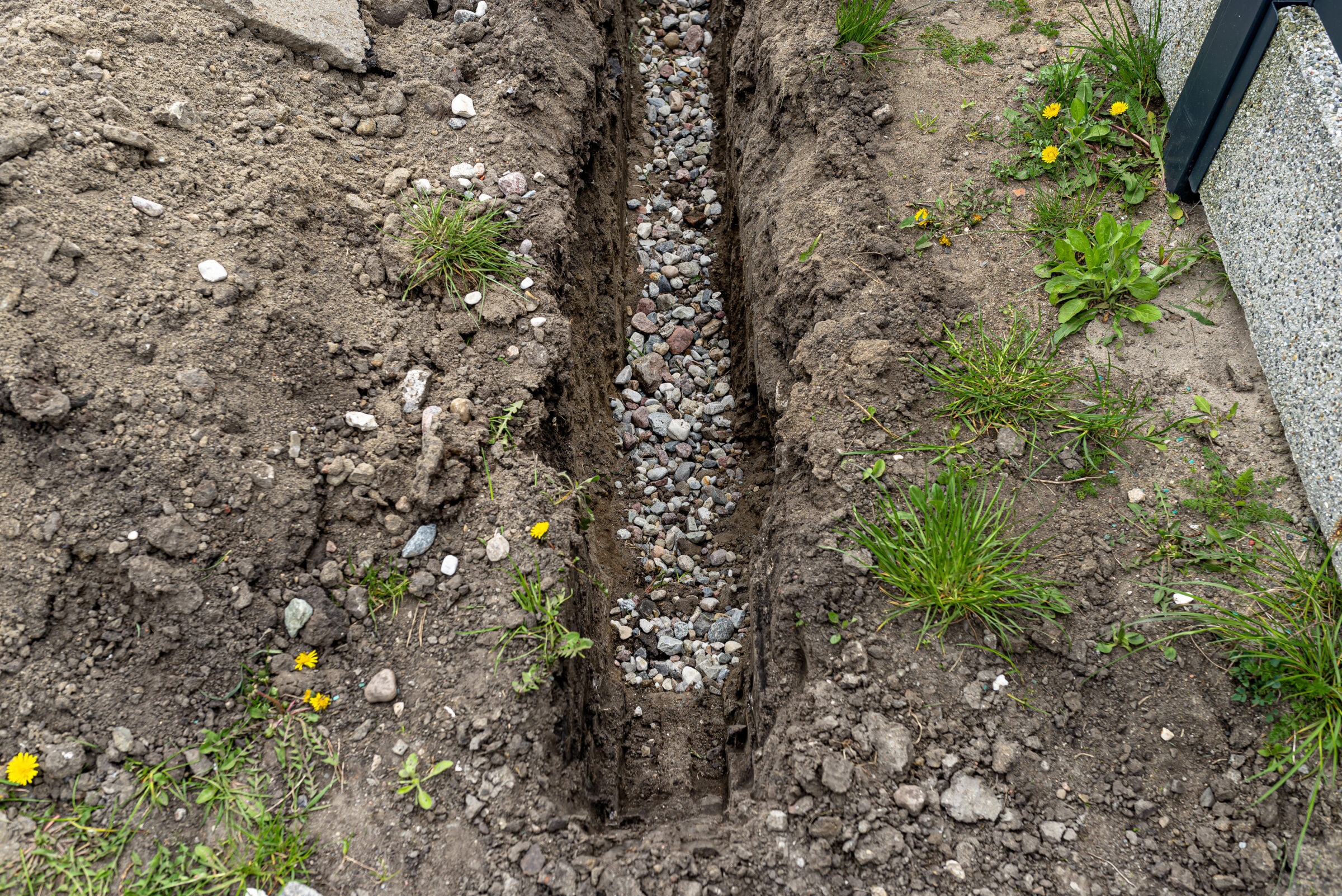 A narrow trench filled with small stones runs through soil and grass, with concrete on the side.