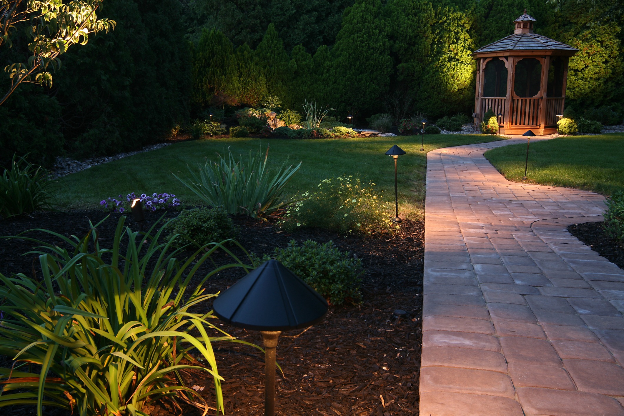 A garden with a brick pathway, surrounded by plants and solar lights, leads to a wooden gazebo at dusk.