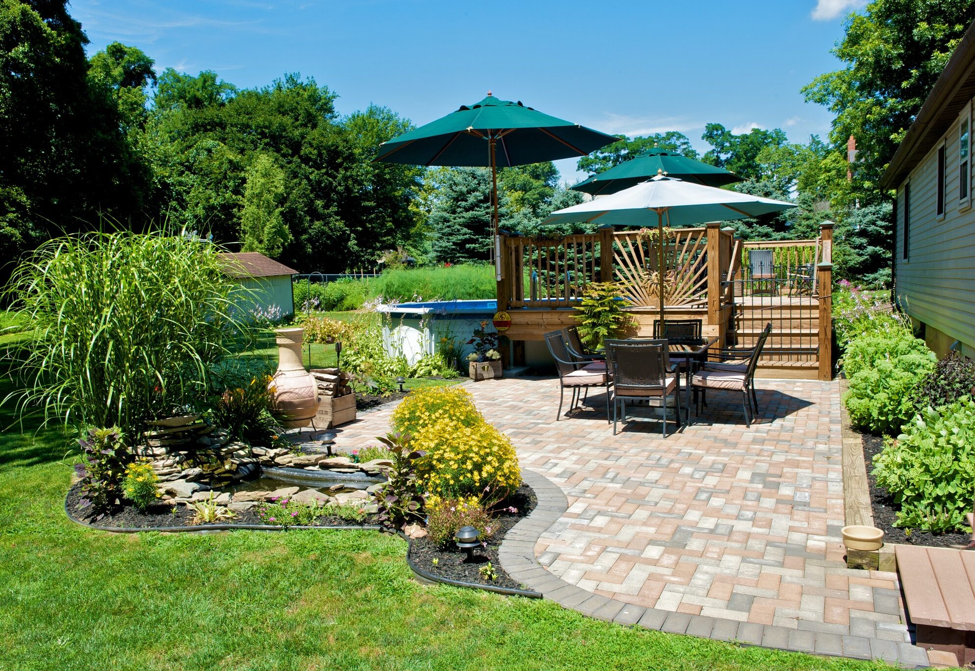 A tranquil backyard with patio furniture, brick paving, umbrellas, lush greenery, small water feature, and a raised deck pool under a clear sky.