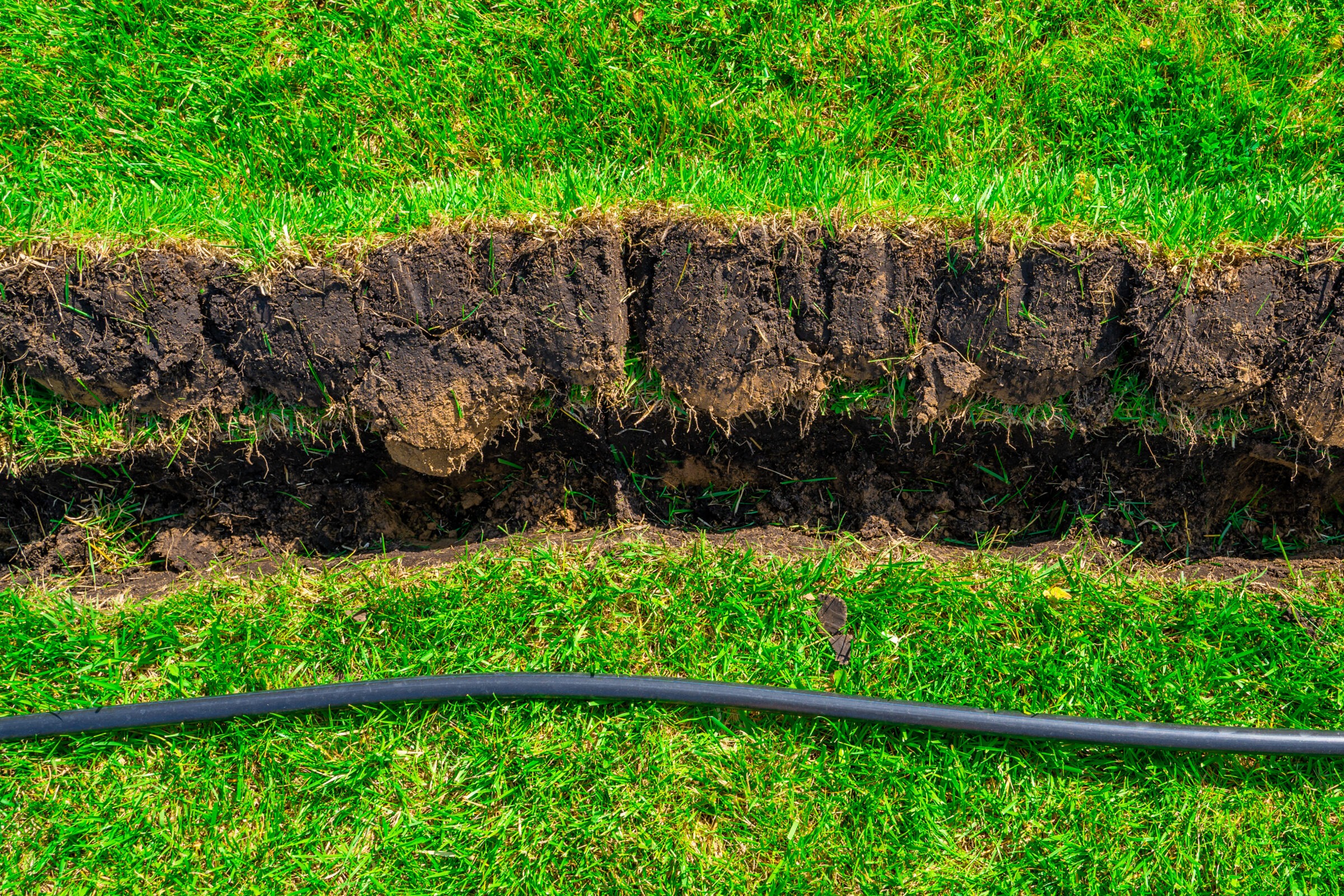 A dug trench in a grassy area with soil exposed, alongside a black cable laid across the vibrant green turf.