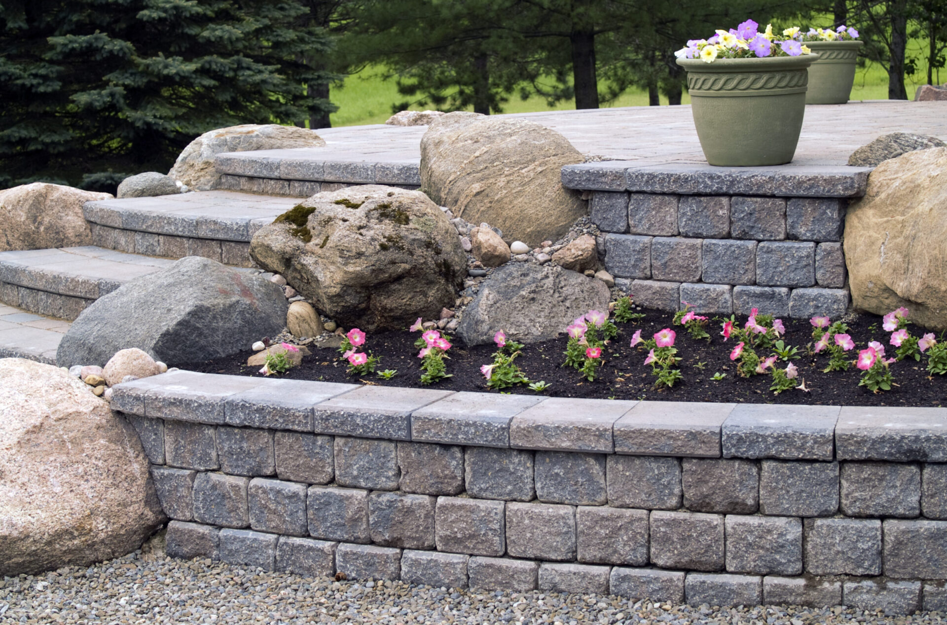 Stone steps with large boulders and colorful flowers, leading to a patio with potted plants. Evergreen trees in the background.