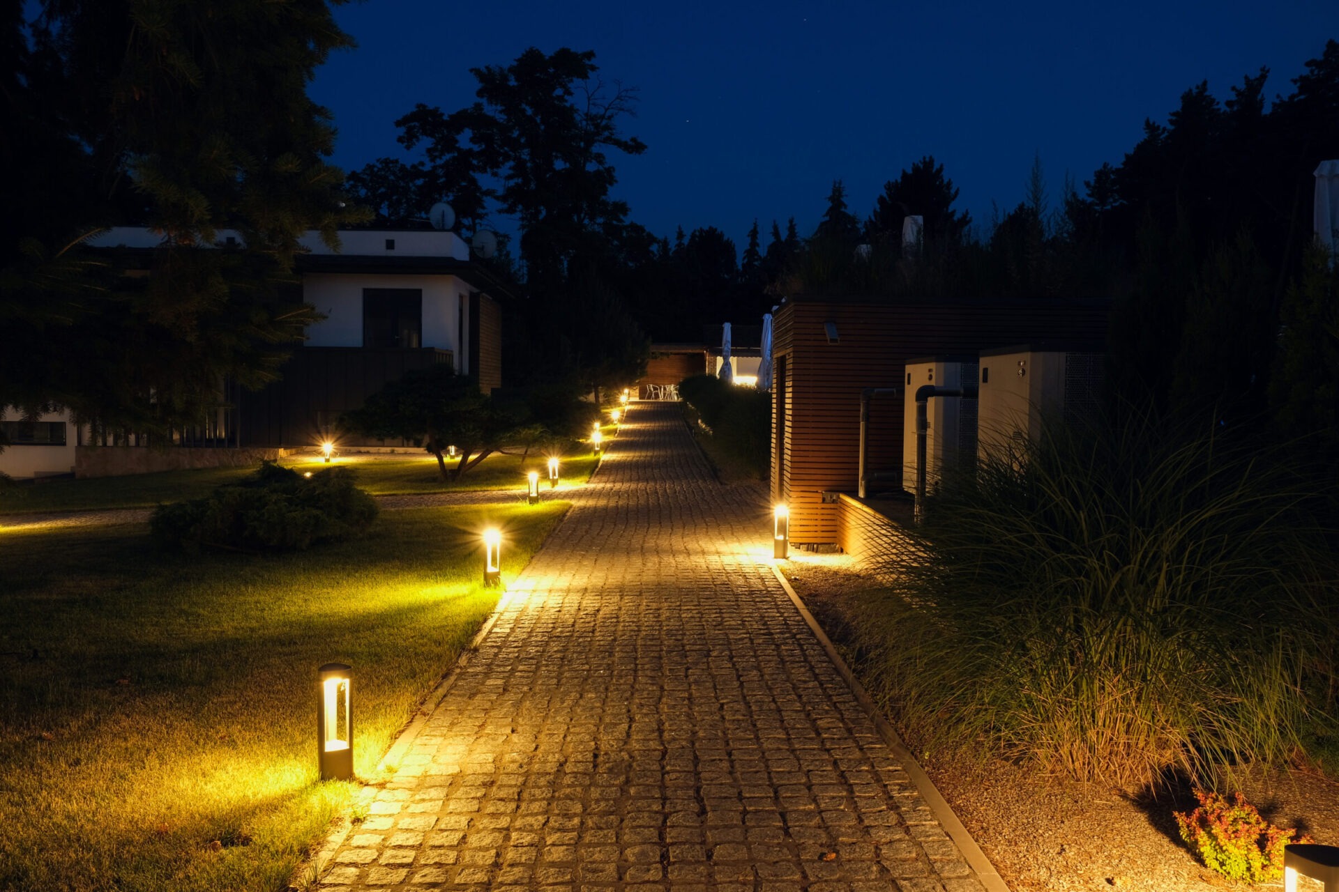 A cobblestone path illuminated by ground lights at night, flanked by modern buildings and trees, creates a serene, inviting atmosphere.