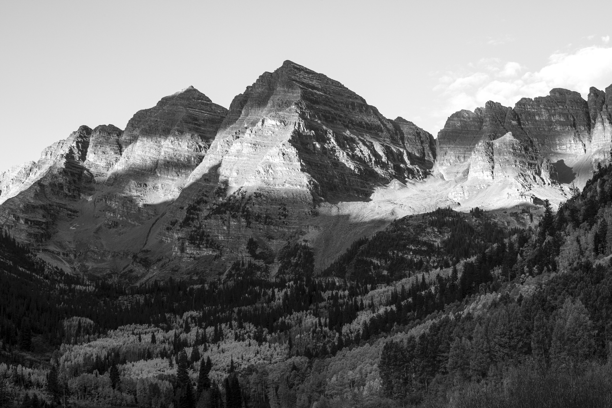 A stunning black and white view of the Maroon Bells mountains, emphasizing shadowed rugged peaks and a dense, expansive forest in the foreground.
