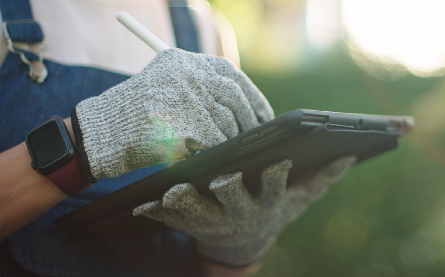 A person wearing gloves and a smartwatch uses a tablet with a stylus in a green, sunny outdoor setting.
