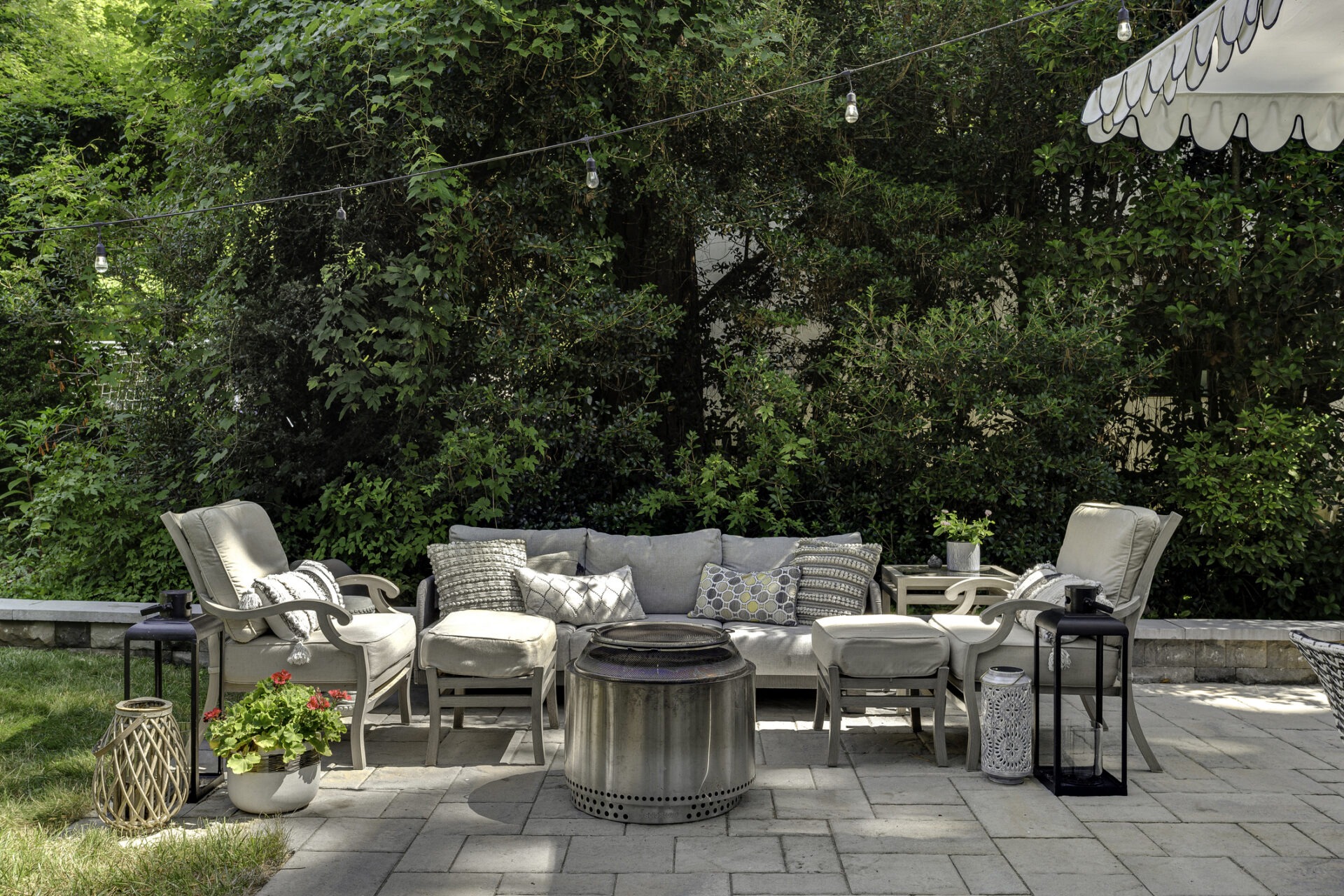 Outdoor patio with cushioned seating, decorative pillows, and a central fire pit. Surrounded by lush greenery and adorned with hanging string lights.