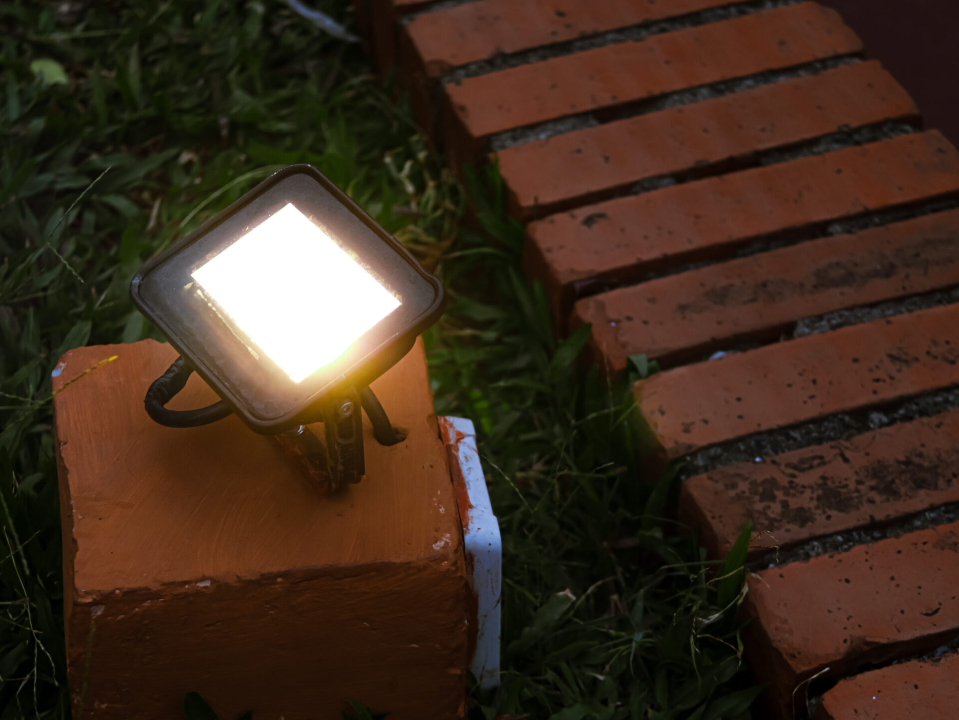 A bright outdoor lamp illuminates a path with red brick border, surrounded by green grass and small plants, creating a serene atmosphere.