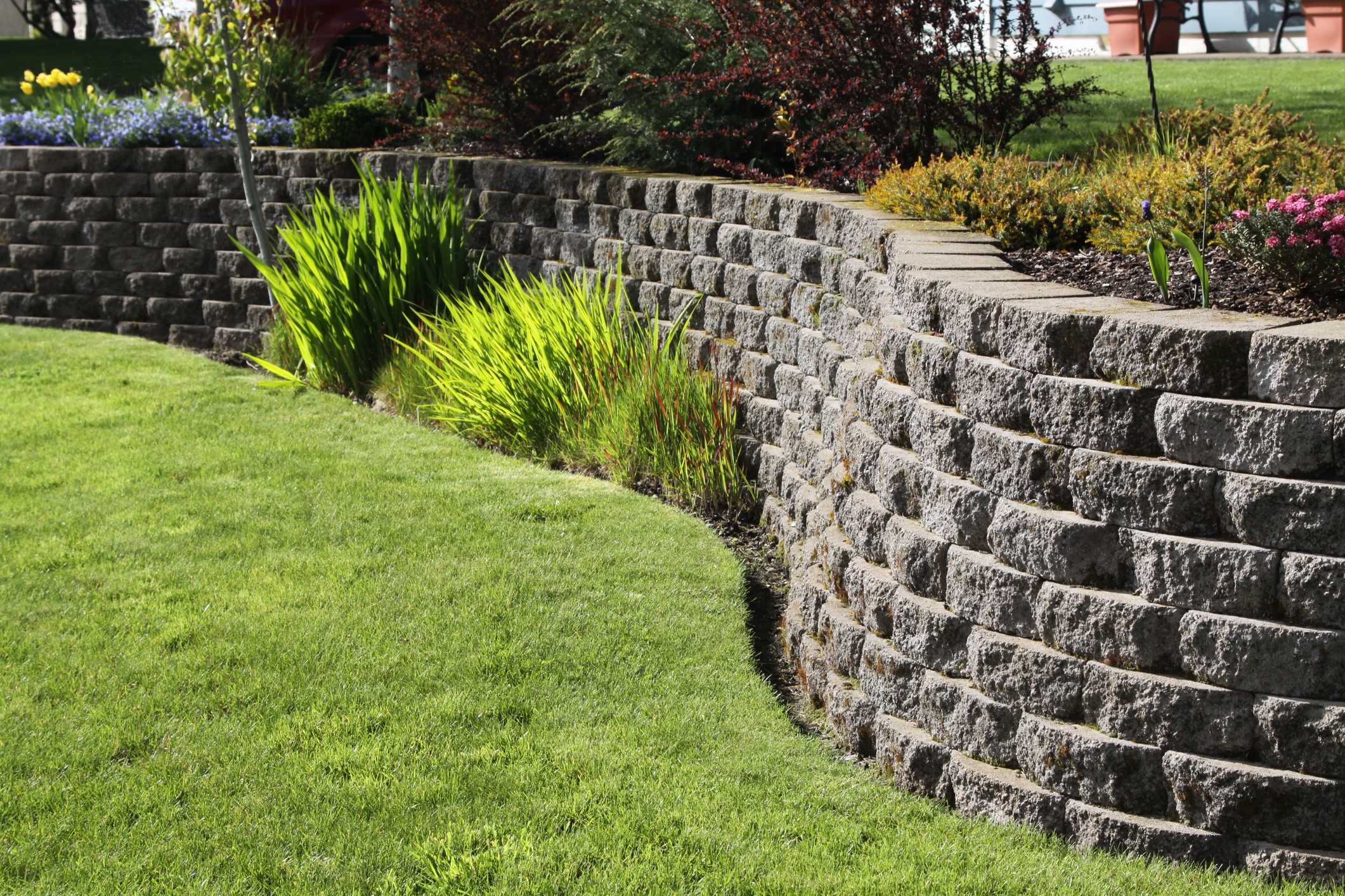 A curved stone retaining wall supports a vibrant garden with lush grass, colorful flowers, and greenery under a sunny sky.