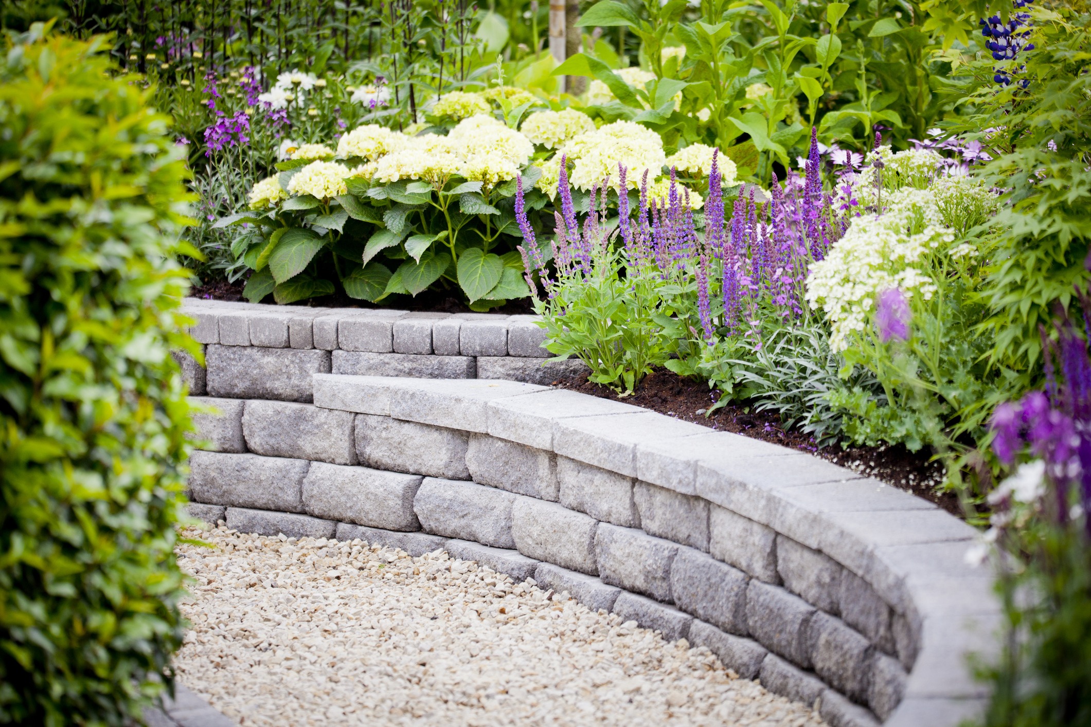 Curved stone wall with vibrant flowers and lush greenery, creating a serene garden atmosphere with a gravel path. No recognizable landmarks present.