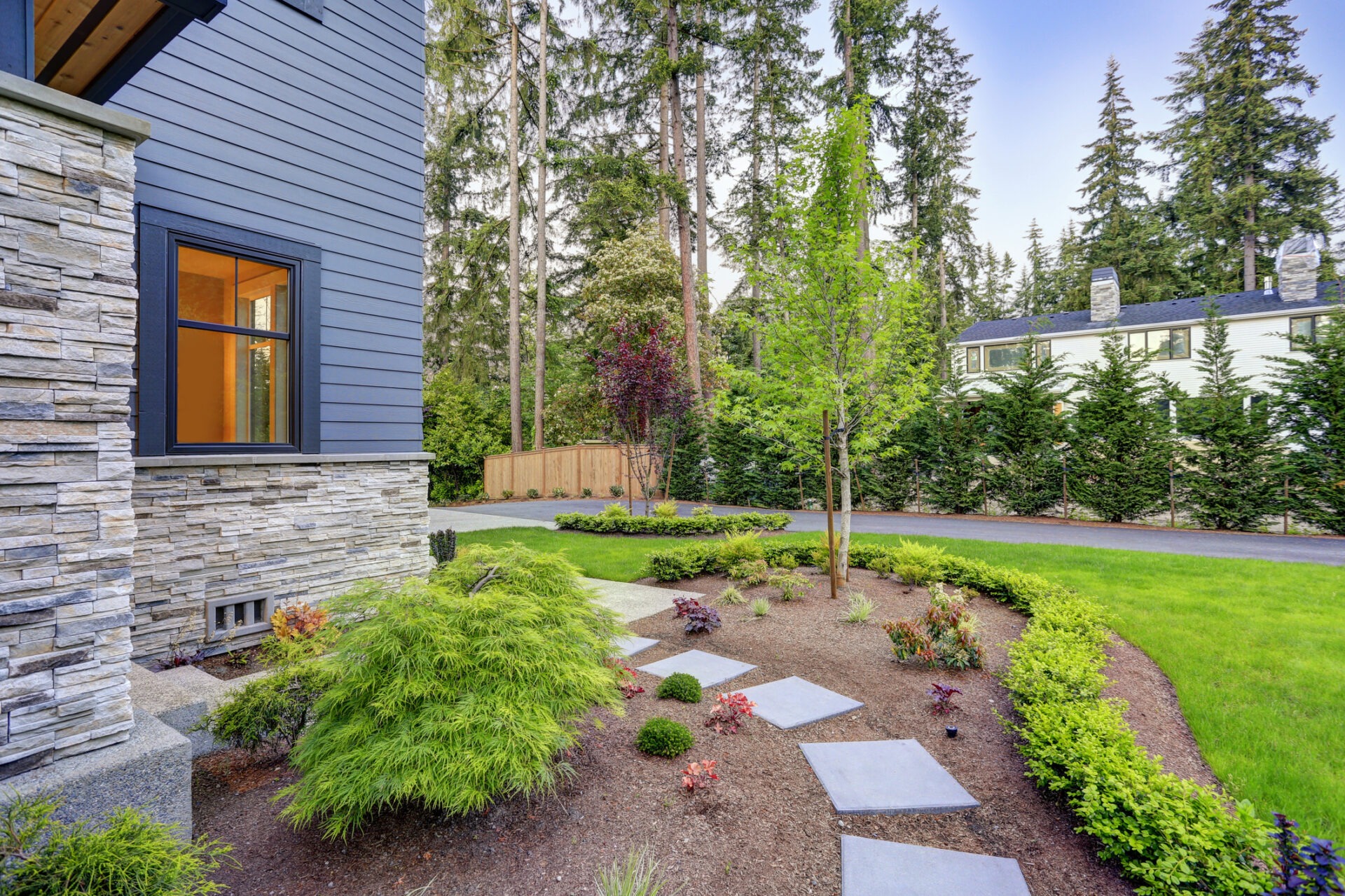 Modern home exterior with stone accents, lush landscaping, and pathway. Surrounded by tall trees and a neatly manicured lawn under a clear sky.