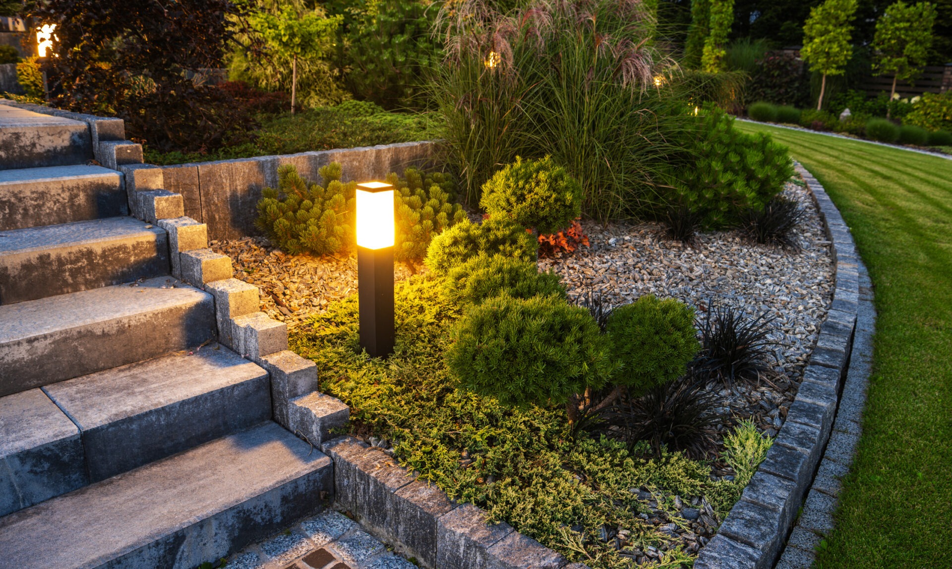 Illuminated garden path with stone steps, bordered by lush greenery and shrubs, featuring a glowing modern pillar light in a landscaped area.