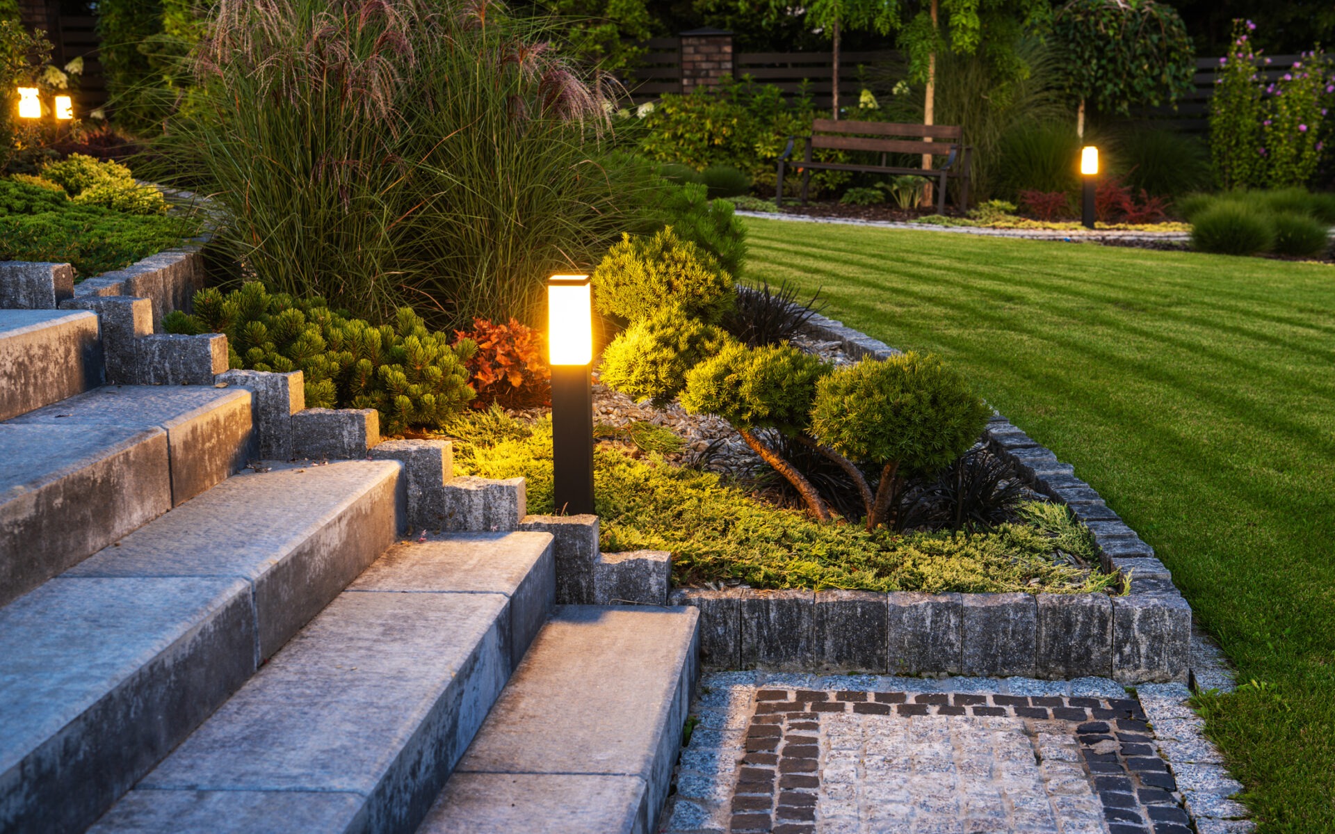 Illuminated garden pathway with stone steps, neatly trimmed bushes, and manicured lawn. Soft lighting enhances the peaceful, landscaped environment at dusk.