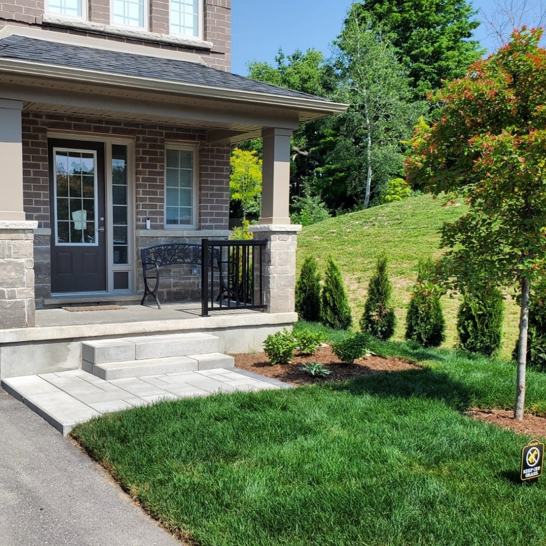 Brick house entrance with porch and a bench, surrounded by well-maintained green lawn and small bushes. Trees in the background.