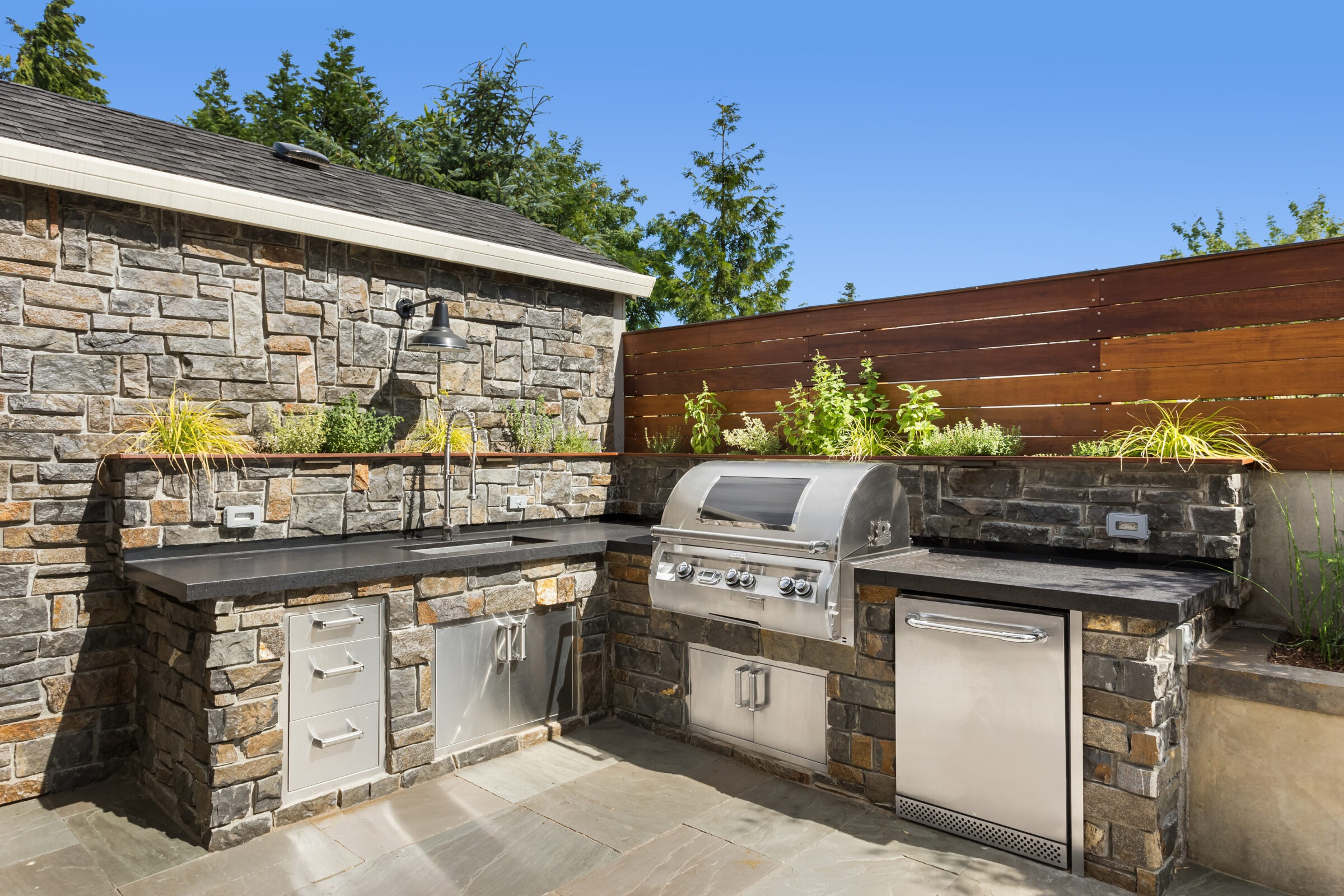 Stone outdoor kitchen with stainless steel appliances, barbecue grill, sink, and herbs in planters. Surrounded by wooden fence and greenery under clear sky.