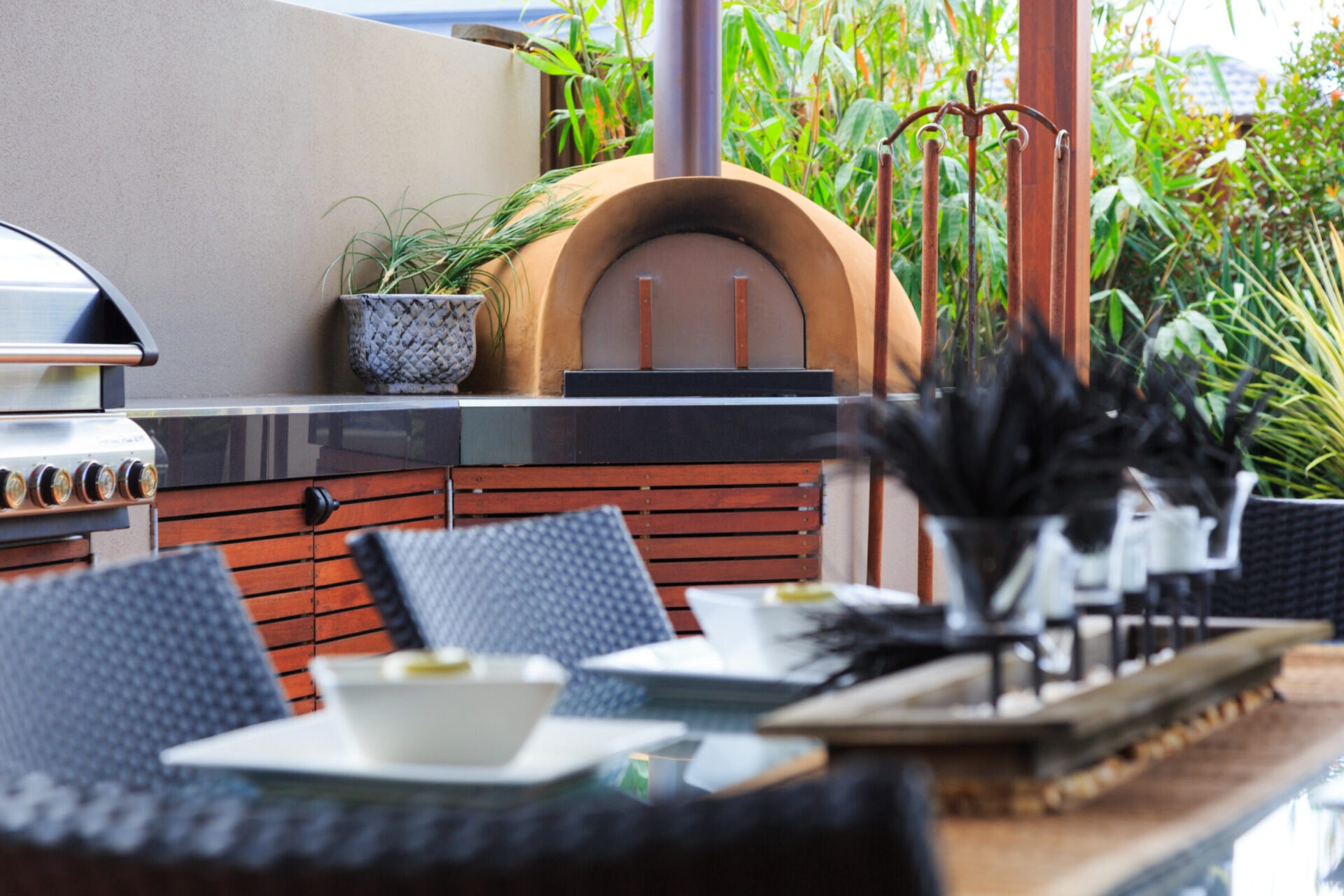 Stylish outdoor patio with wood-fired pizza oven, grill, wicker chairs, table set with bowls, surrounded by lush green plants. Cozy and inviting ambiance.